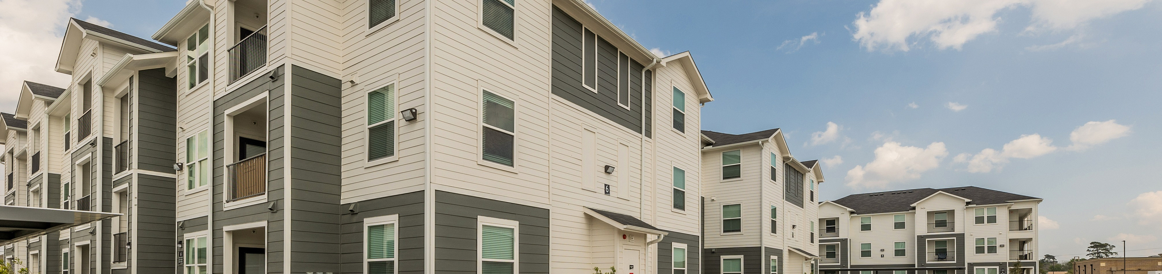 Charming modern apartment complex featuring multi-level buildings with balconies, surrounded by ample parking and manicured landscaping under a clear blue sky.