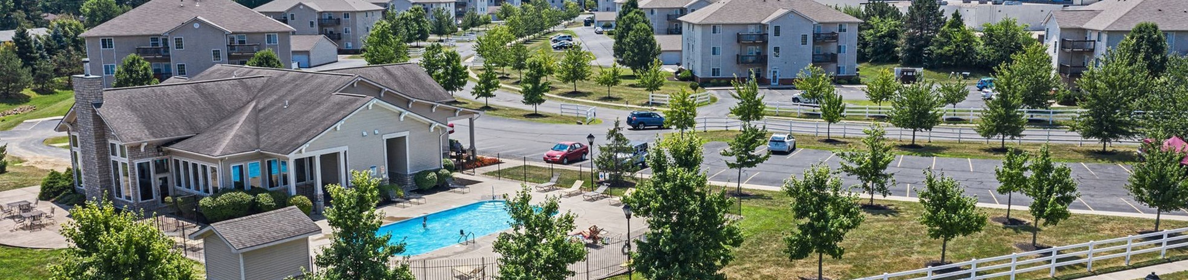 Scenic aerial view of a modern apartment complex with landscaped grounds, a sparkling swimming pool, and ample parking, nestled in a serene neighborhood.
