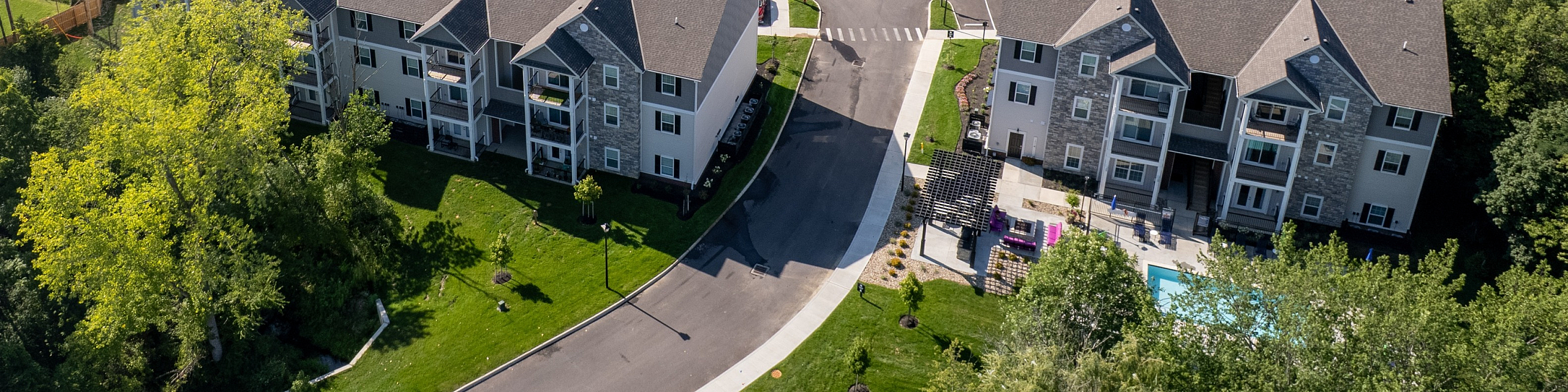 Aerial view of modern apartment complex surrounded by lush greenery and open landscapes, featuring multiple buildings with ample parking, offering a serene living experience.