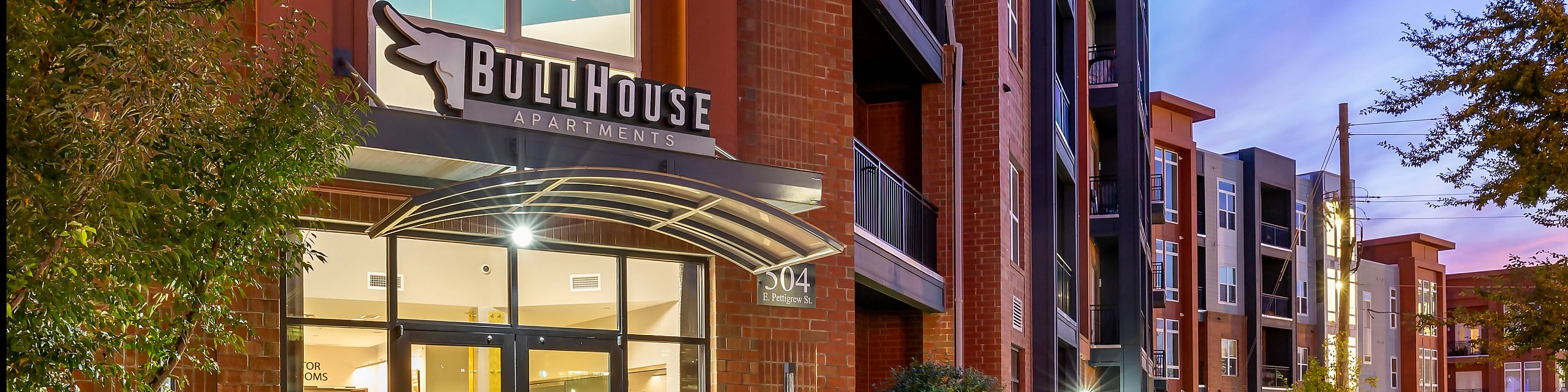 Inviting entrance of modern BullHouse Apartments, surrounded by colorful planters and stylish architecture during twilight.