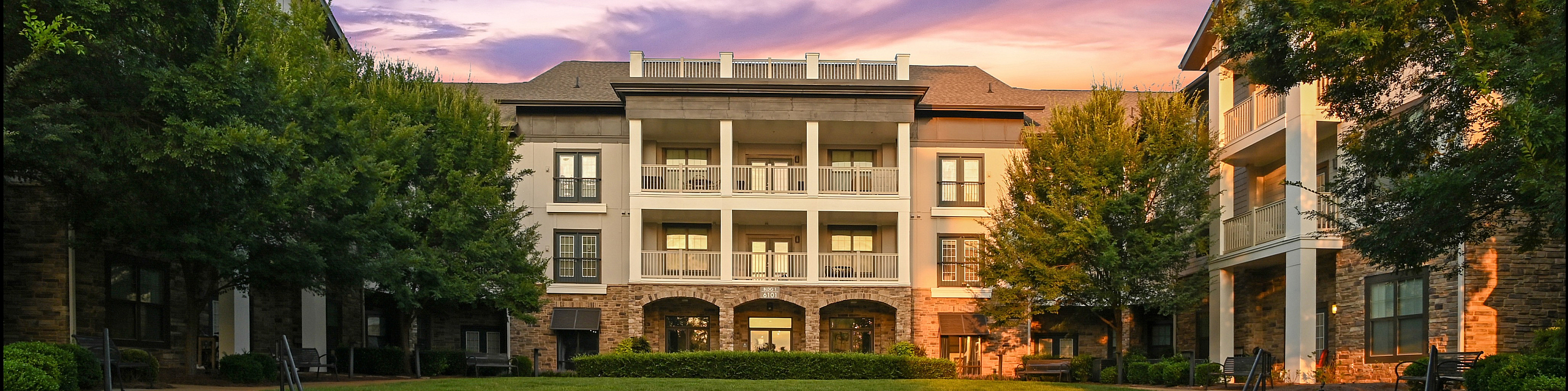 Elegant apartment building with lush landscaping and a serene sunset backdrop, promising a tranquil and luxurious living experience.