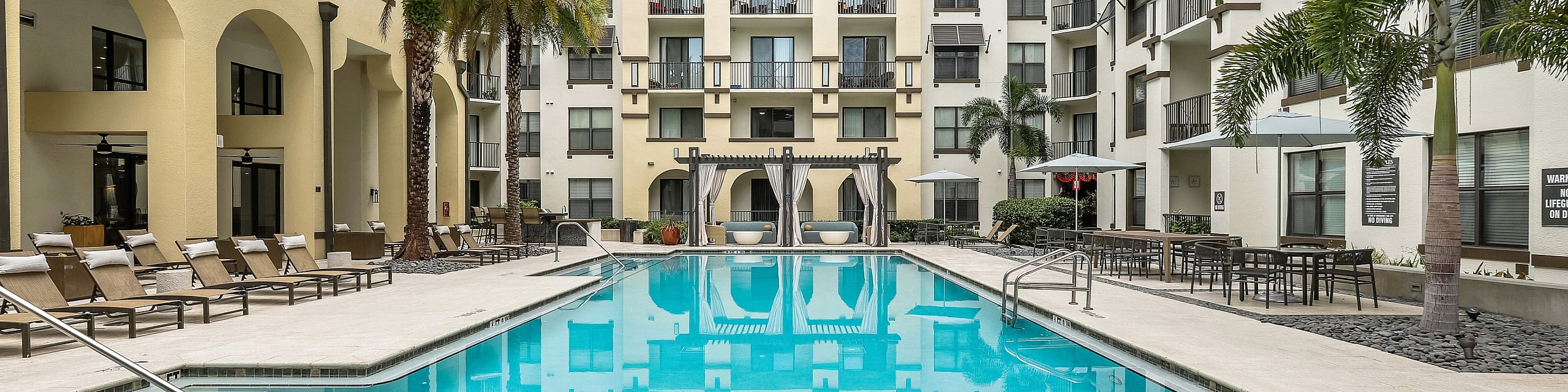 Luxurious apartment complex with a sparkling outdoor pool, surrounded by stylish lounge chairs, palm trees, and elegant architecture.
