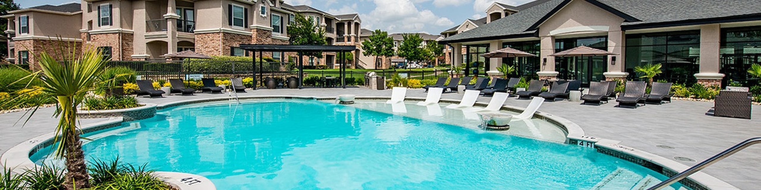 Luxurious apartment complex featuring a stunning pool area with sun loungers, lush landscaping, and beautifully designed buildings under a clear blue sky.