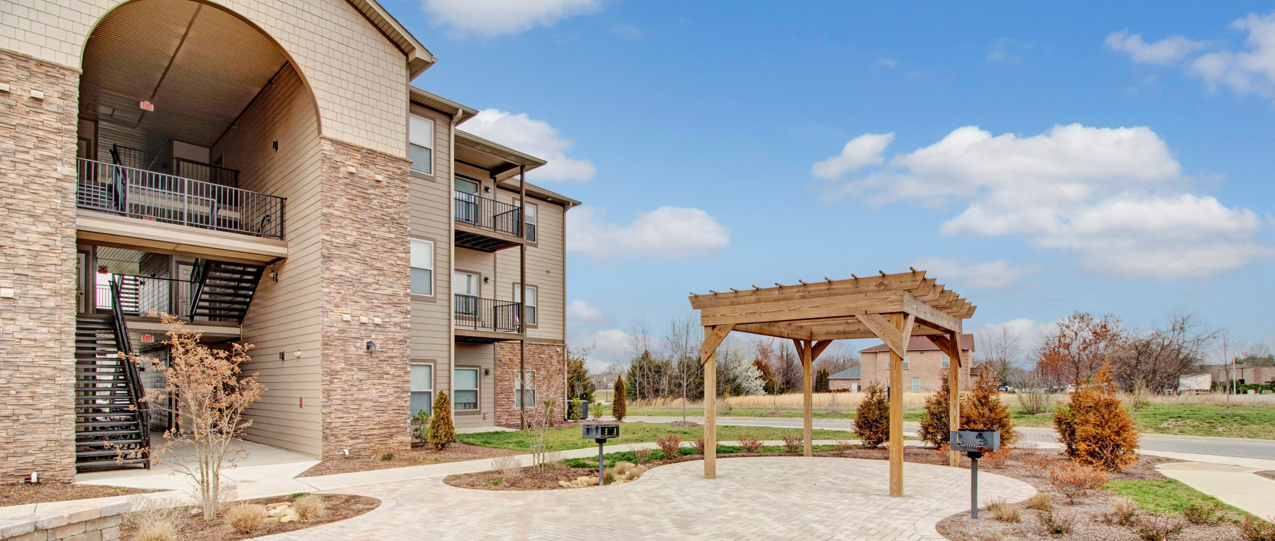 Part of the Merchants Walk Apartment building with stone and wood siding with brick walkway and wood pergolas with grass around it and road in background