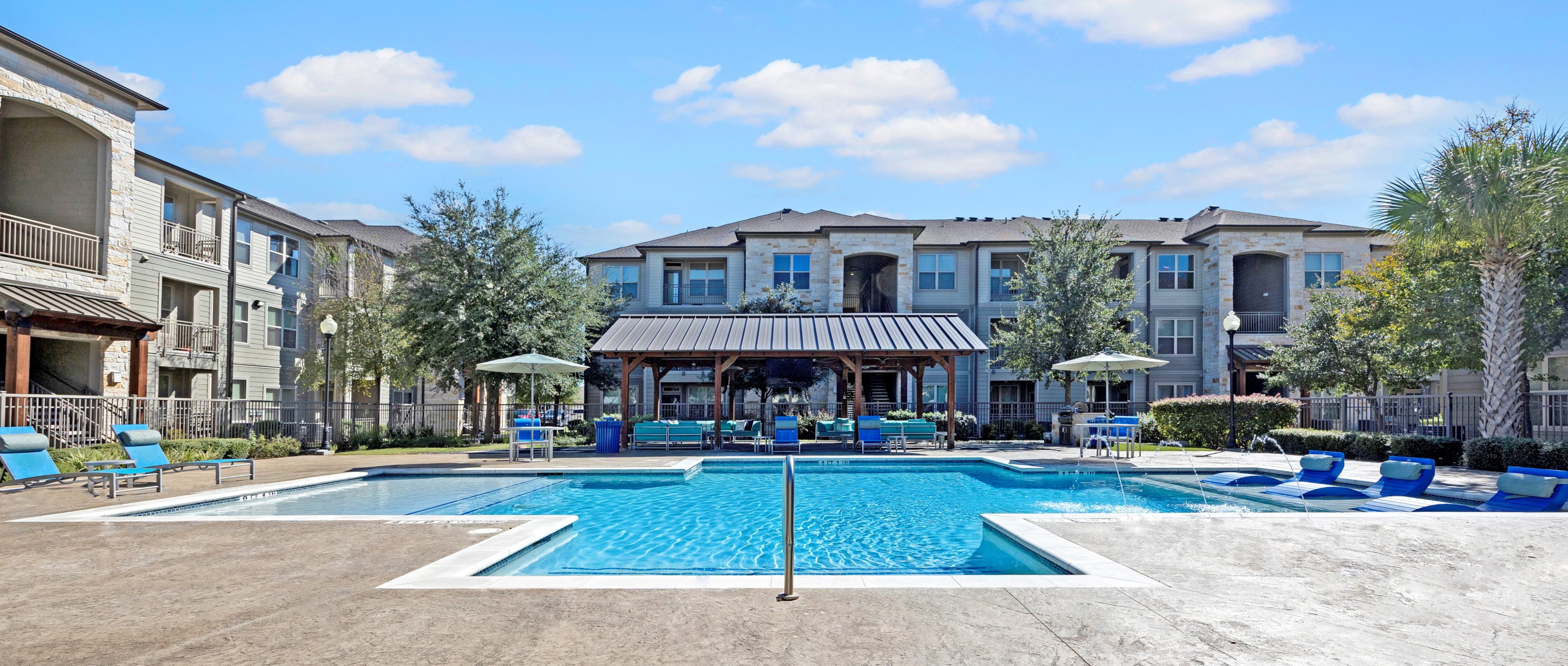 Swimming pool area with blue chairs around it and white fence around pool with wood covering in the back area of pool with Oaks of Kyle apartment building behind