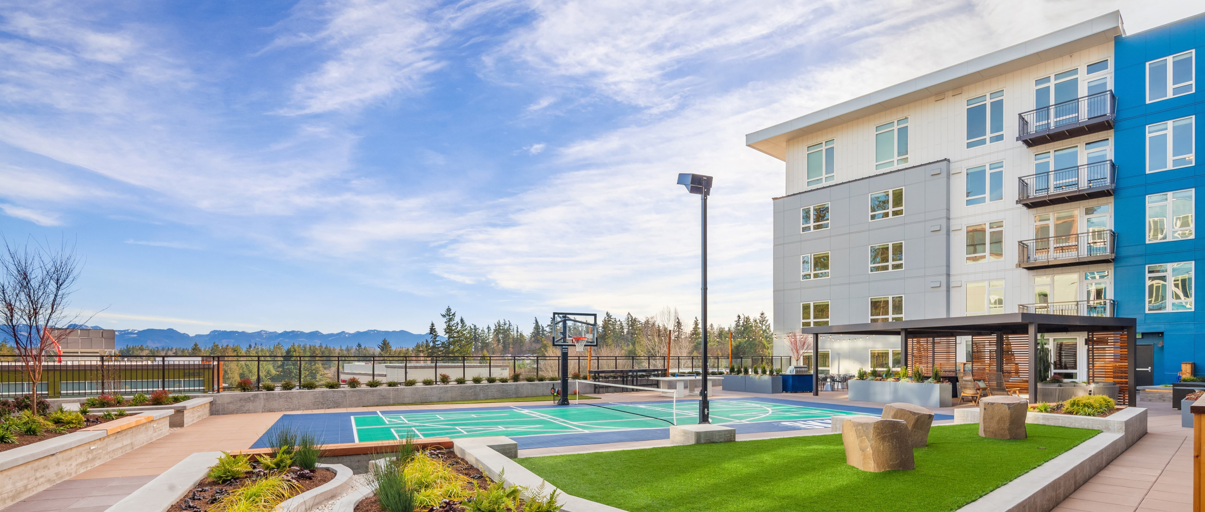 Concrete paths and grass patch with basketball court and blue, gray and white SKY apartment building with trees and mountains in the background