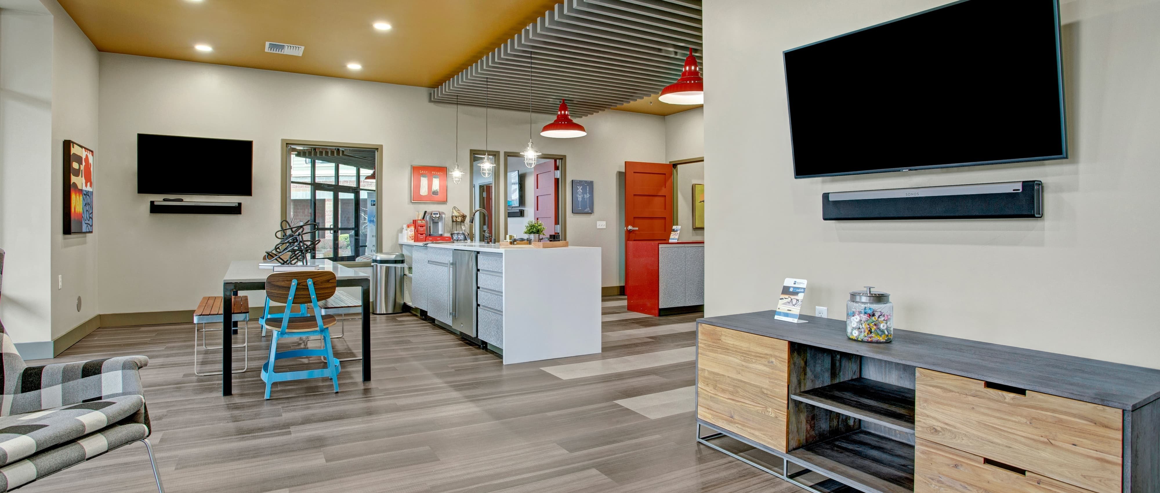 Leasing Office With Blue Chair and Bench at at table with wood entertainment stand and and counter top with coffee supplies and white walls with two TVs