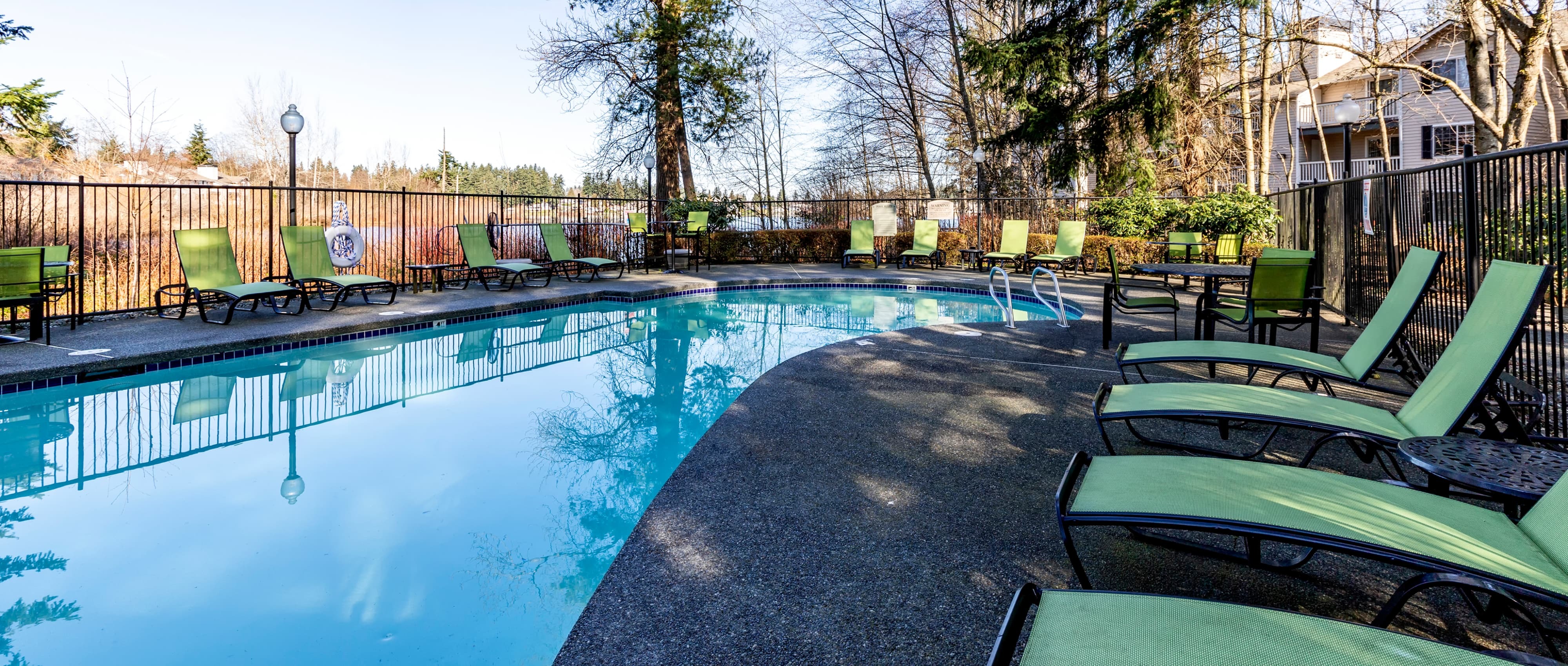 Swimming pool and pool area with green pool chairs and black metal fence around pool area with trees and black metal poll light and cream colored apartment build of Charter Club Apartments
