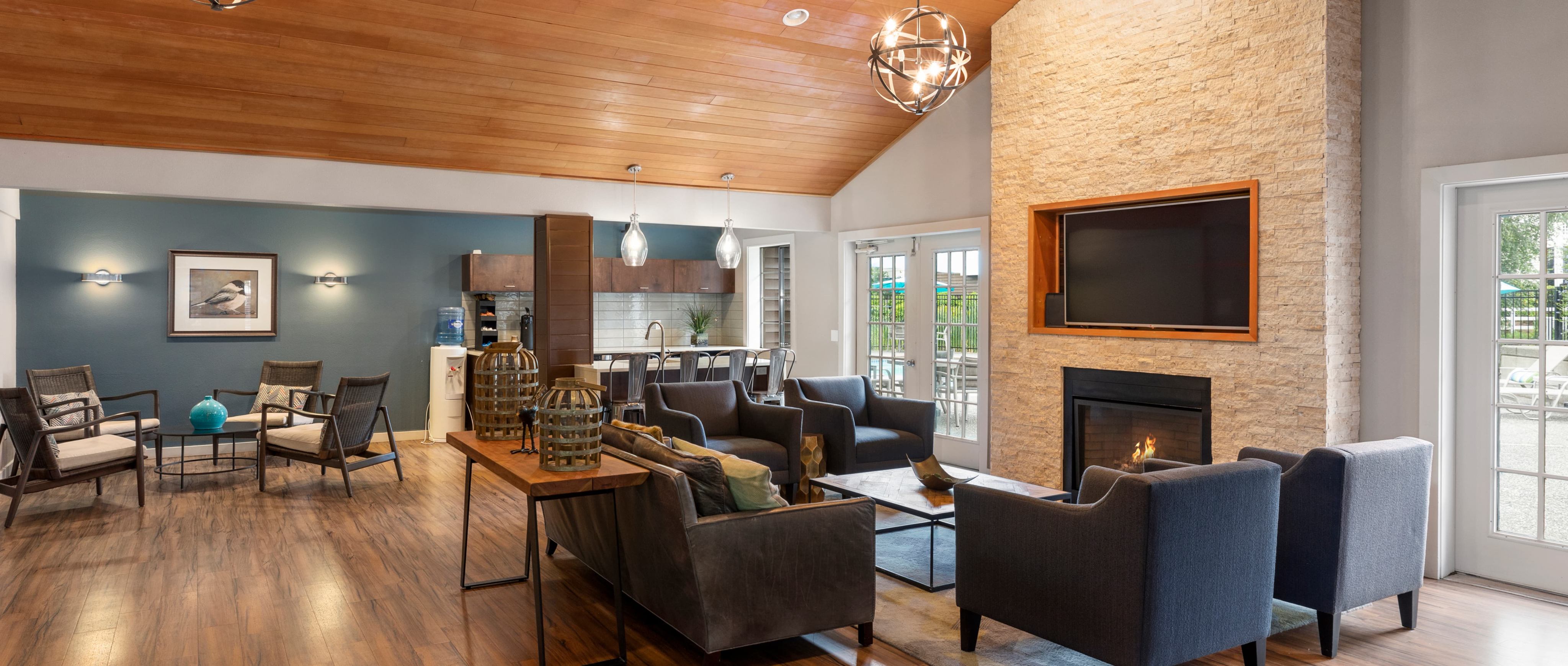 Clubhouse at Olin Fields Apartments with gray fabric chairs and couch around stone fireplace with wood framed TV above and chairs around coffee table and bar area behind