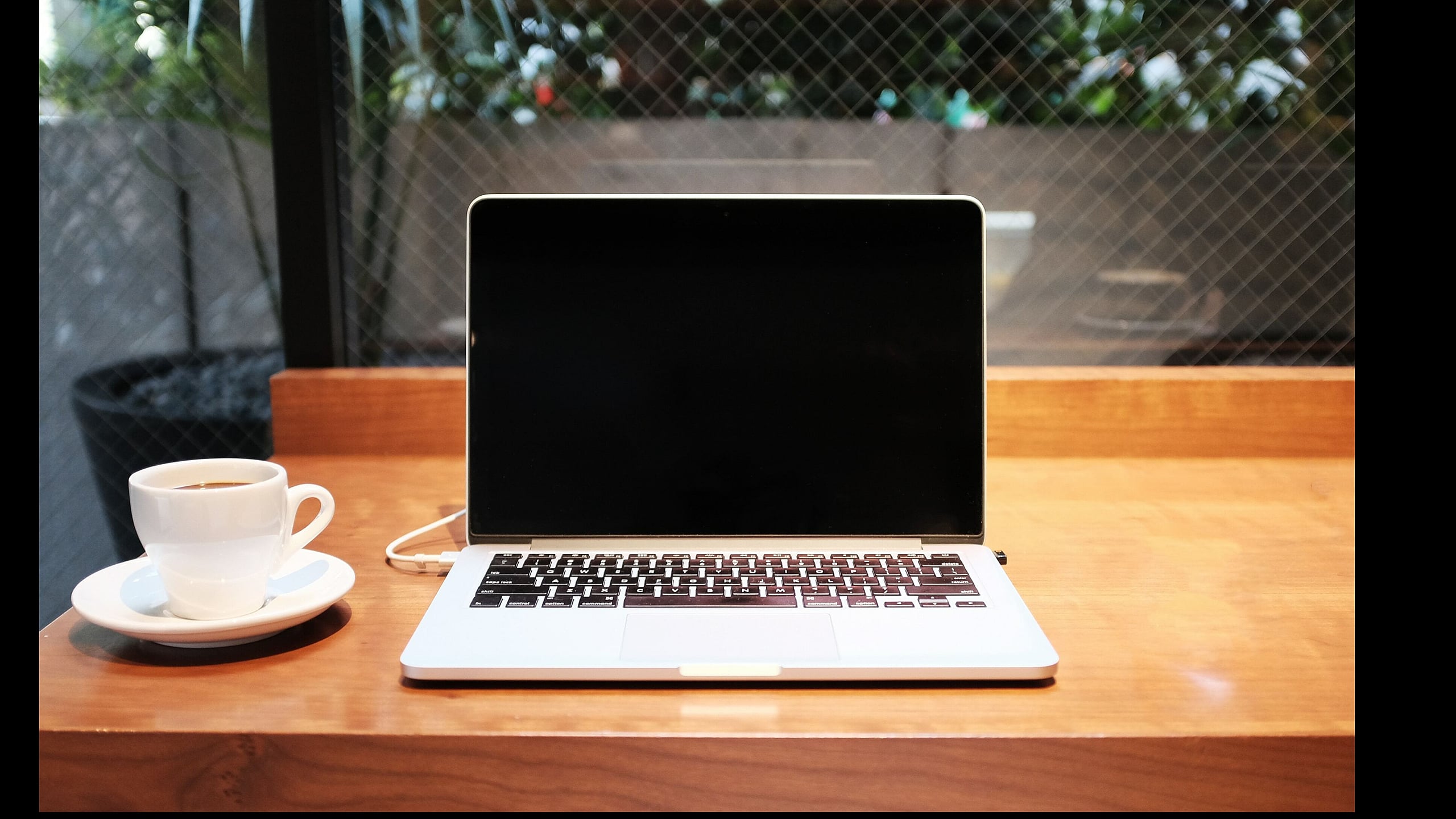 Cozy workspace with an elegant laptop and steaming cup of coffee on a wooden table by a large window, offering a serene view.