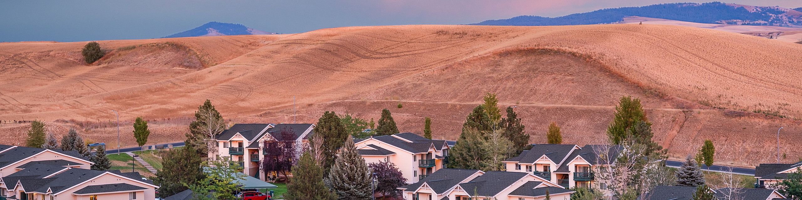 Charming apartment complex nestled in a picturesque landscape with rolling hills and a vibrant sunset sky.