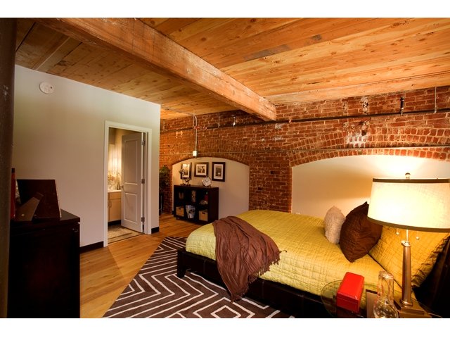 Cozy bedroom with bed tucked against exposed brick wall. Exposed beam ceiling and hardwood flooring with en suite bathroom.