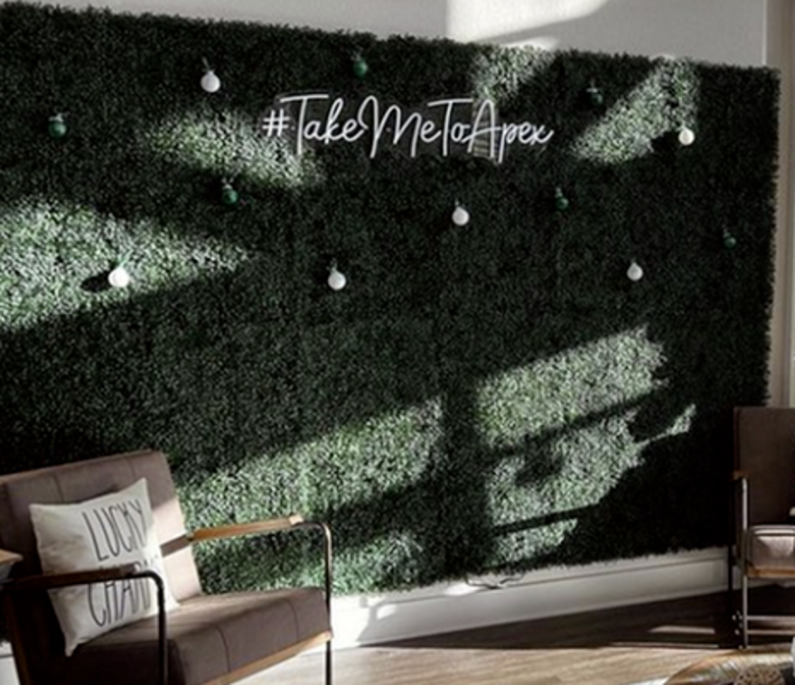 Green foliage wall with a neon sign saying #Take Me To Apex