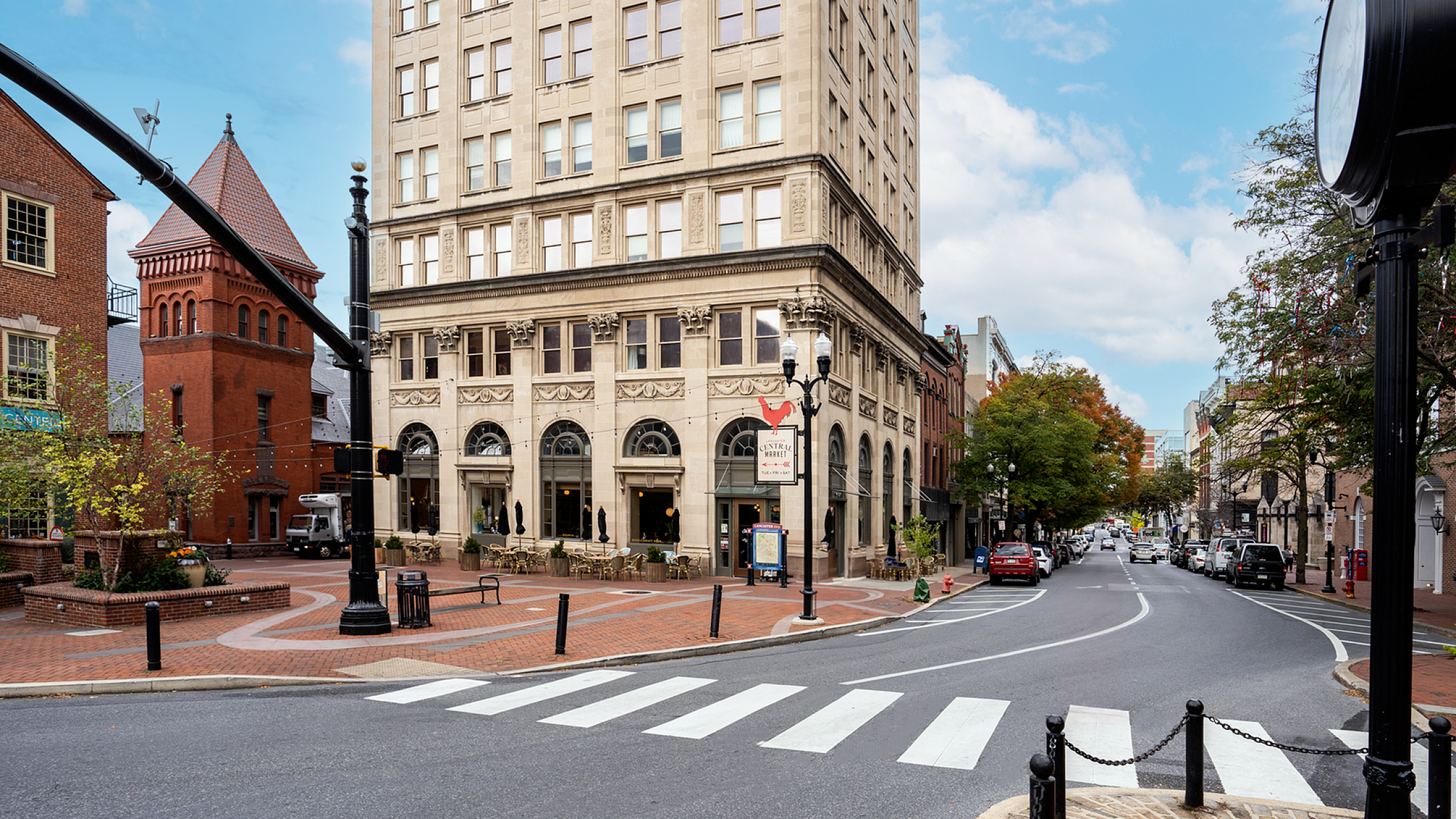 Charming urban street view with classic architecture, featuring a grand historic building with arched windows, nestled in a lively cityscape with vibrant greenery and quaint outdoor seating.