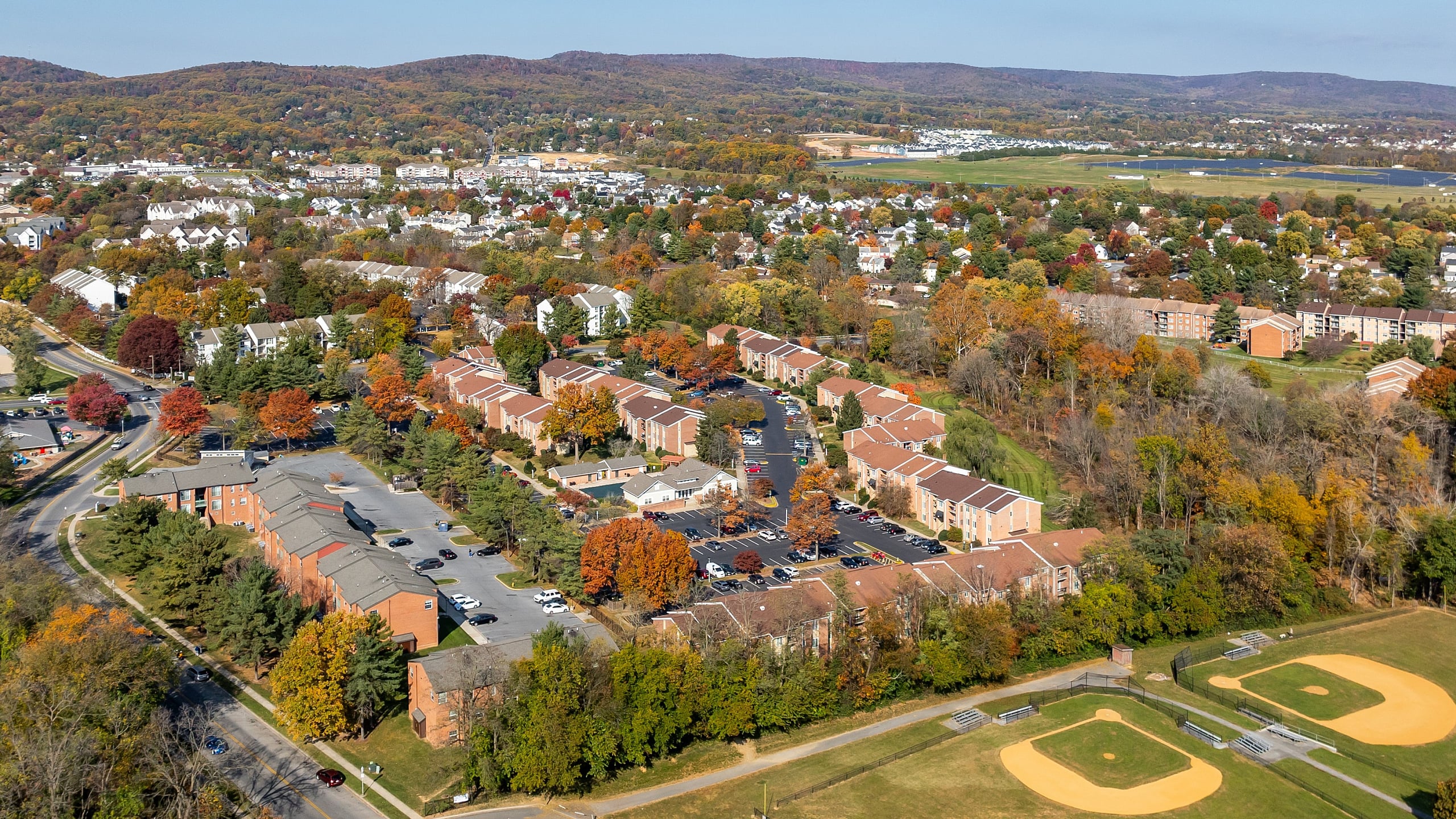 Charming aerial view of a vibrant residential community surrounded by lush greenery and rolling hills, featuring spacious townhomes and well-maintained sports fields.