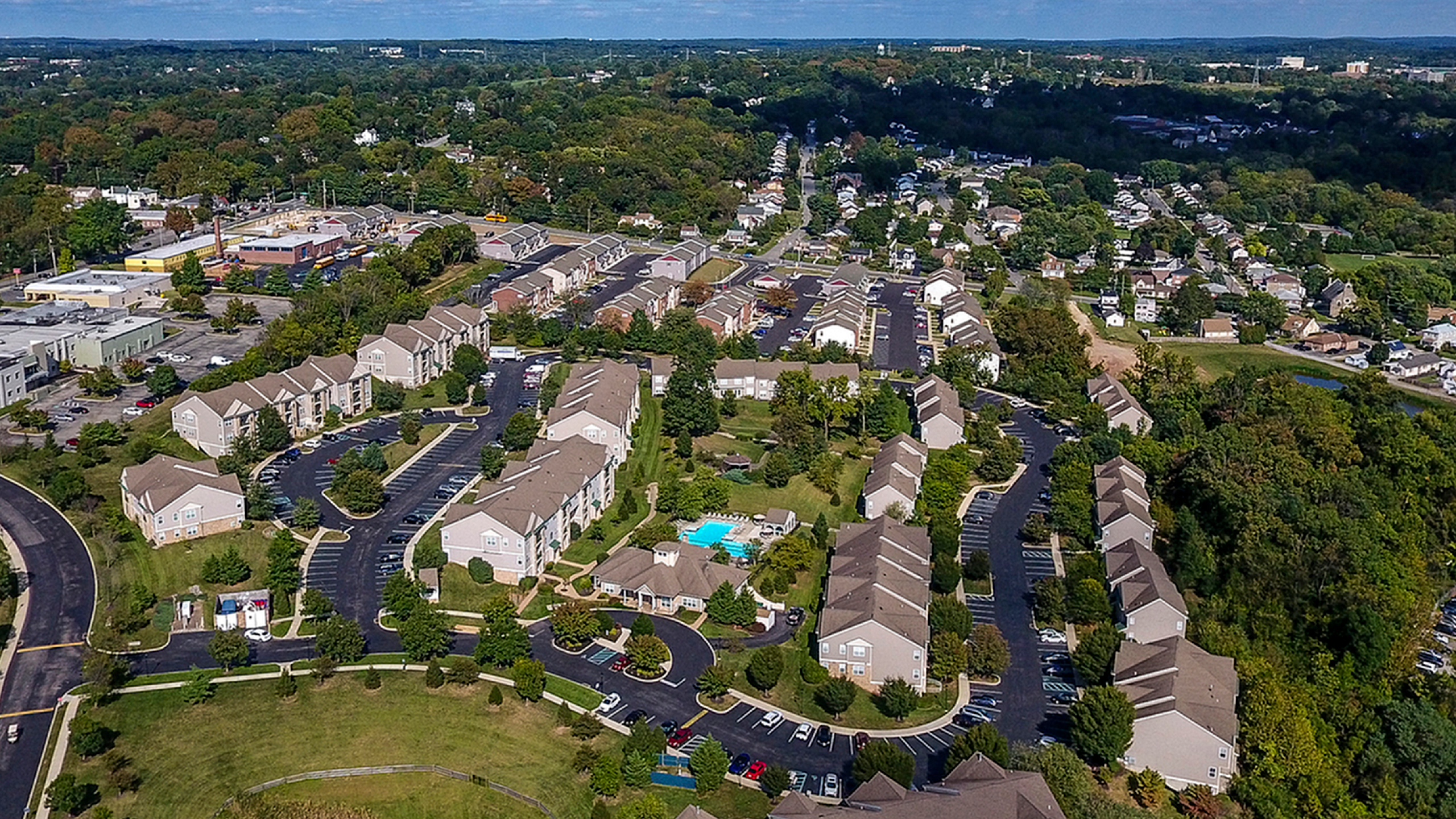 Aerial view of a picturesque apartment complex surrounded by lush greenery, featuring well-planned roads, modern buildings, and a sparkling blue pool, offering a serene and inviting community atmosphere.