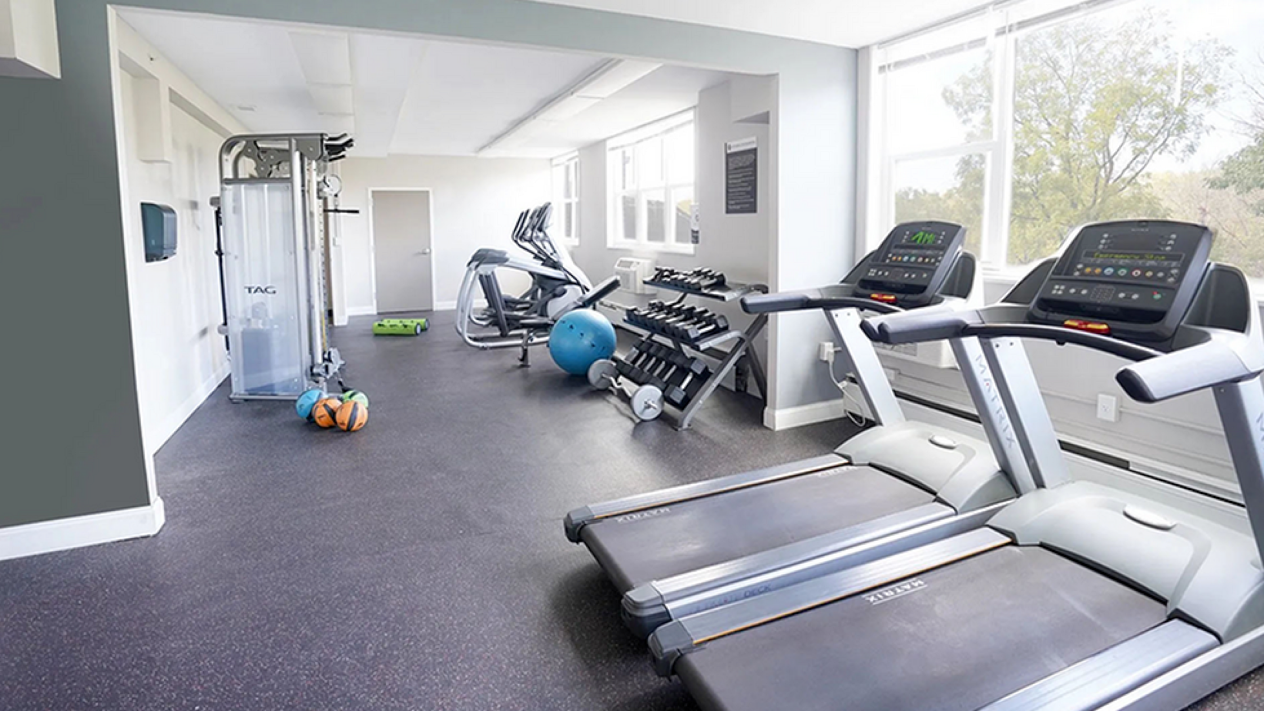 Bright, modern gym featuring treadmills, exercise bikes, and a variety of fitness equipment in a spacious setting with large windows.