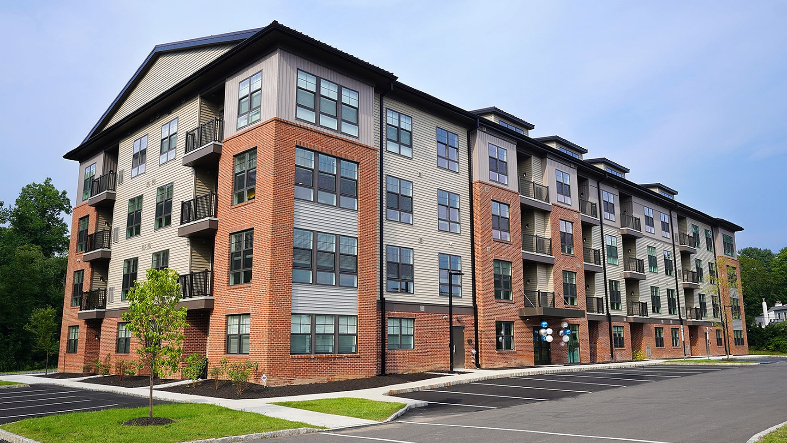 Modern four-story apartment building featuring stylish brick and siding facade with spacious balconies and ample parking, surrounded by lush greenery.