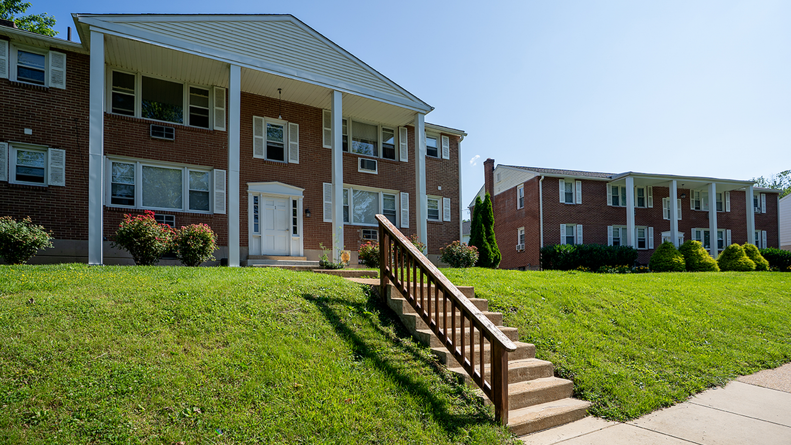 Charming red brick apartment buildings with classic white pillars and lush green lawns under a clear blue sky.