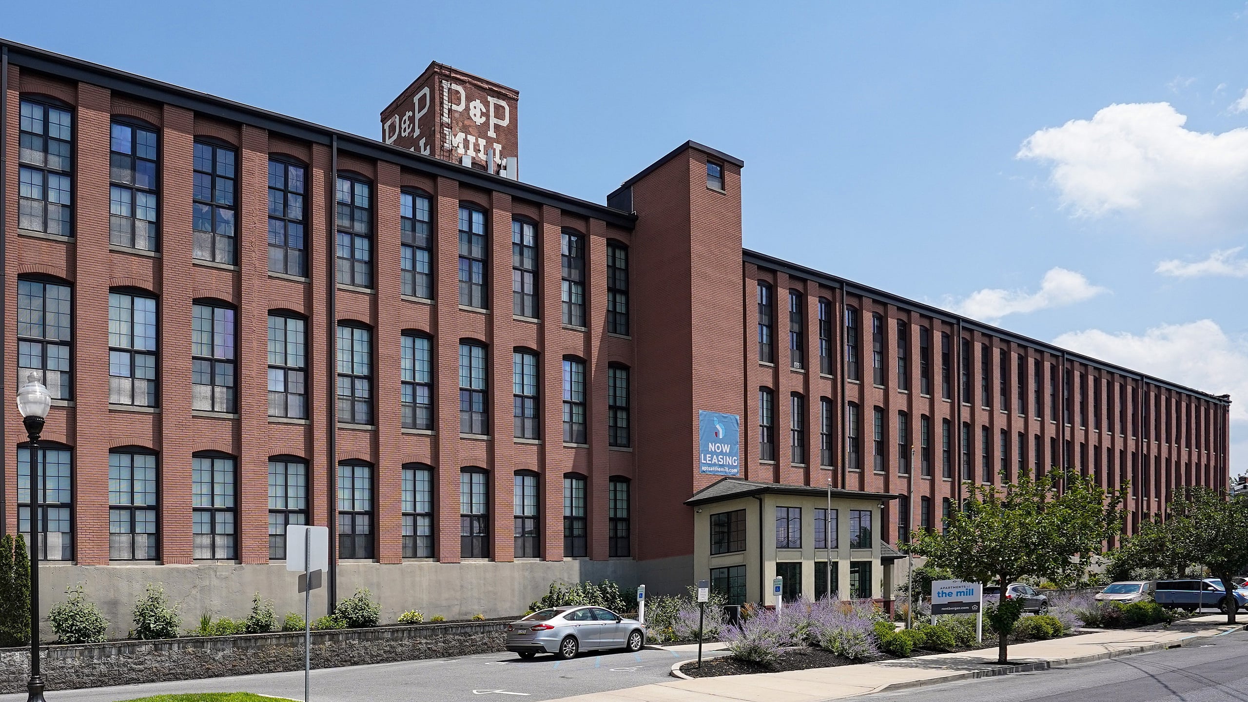 Historic red-brick building with large windows offering modern loft apartments, now leasing, in a vibrant urban setting.