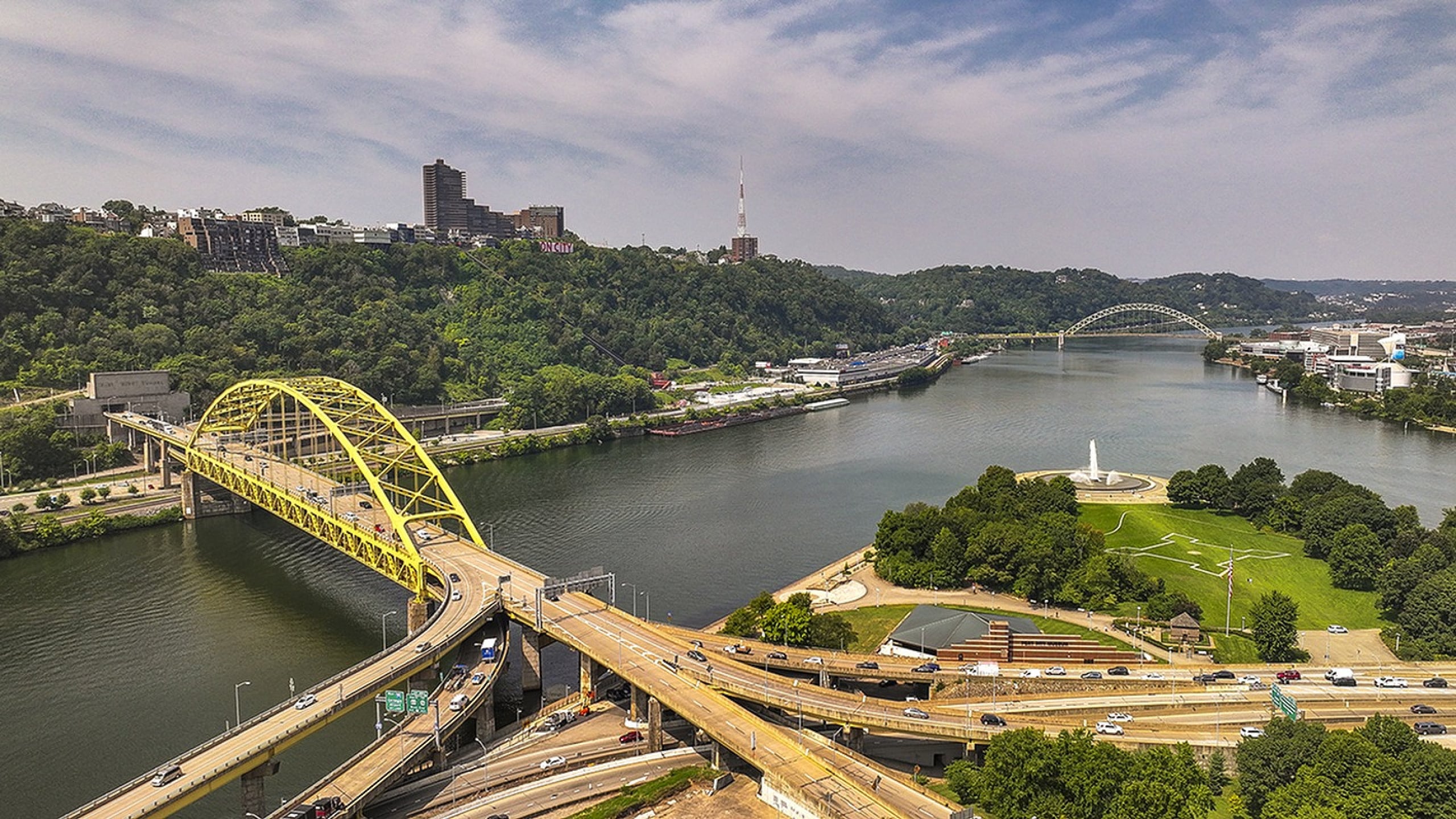 Breathtaking aerial view of a vibrant cityscape featuring a striking yellow bridge spanning a serene river, surrounded by lush greenery and urban architecture.