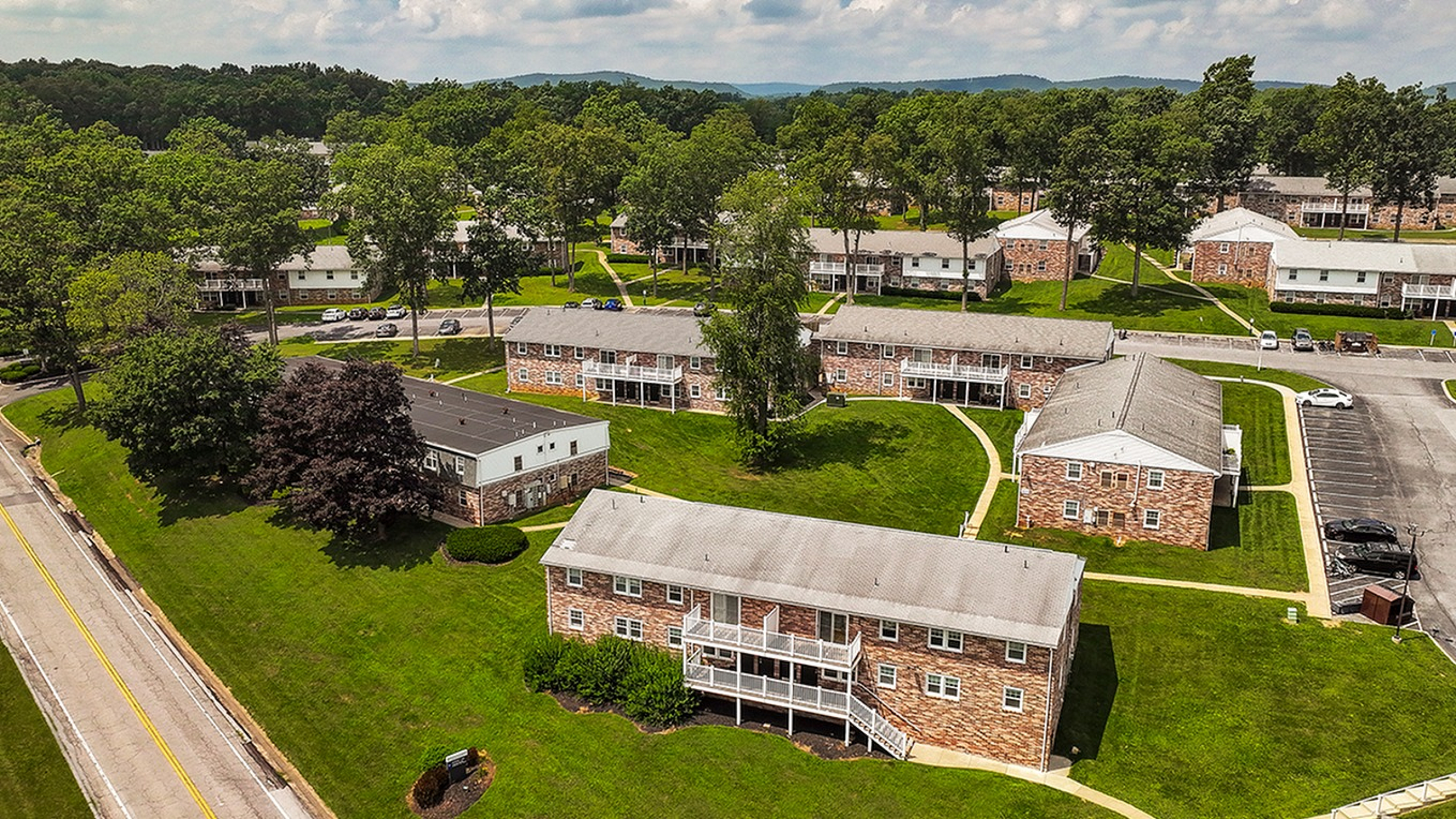 Charming aerial view of a picturesque apartment complex with brick buildings, lush green lawns, and surrounding trees creating a serene and welcoming environment.