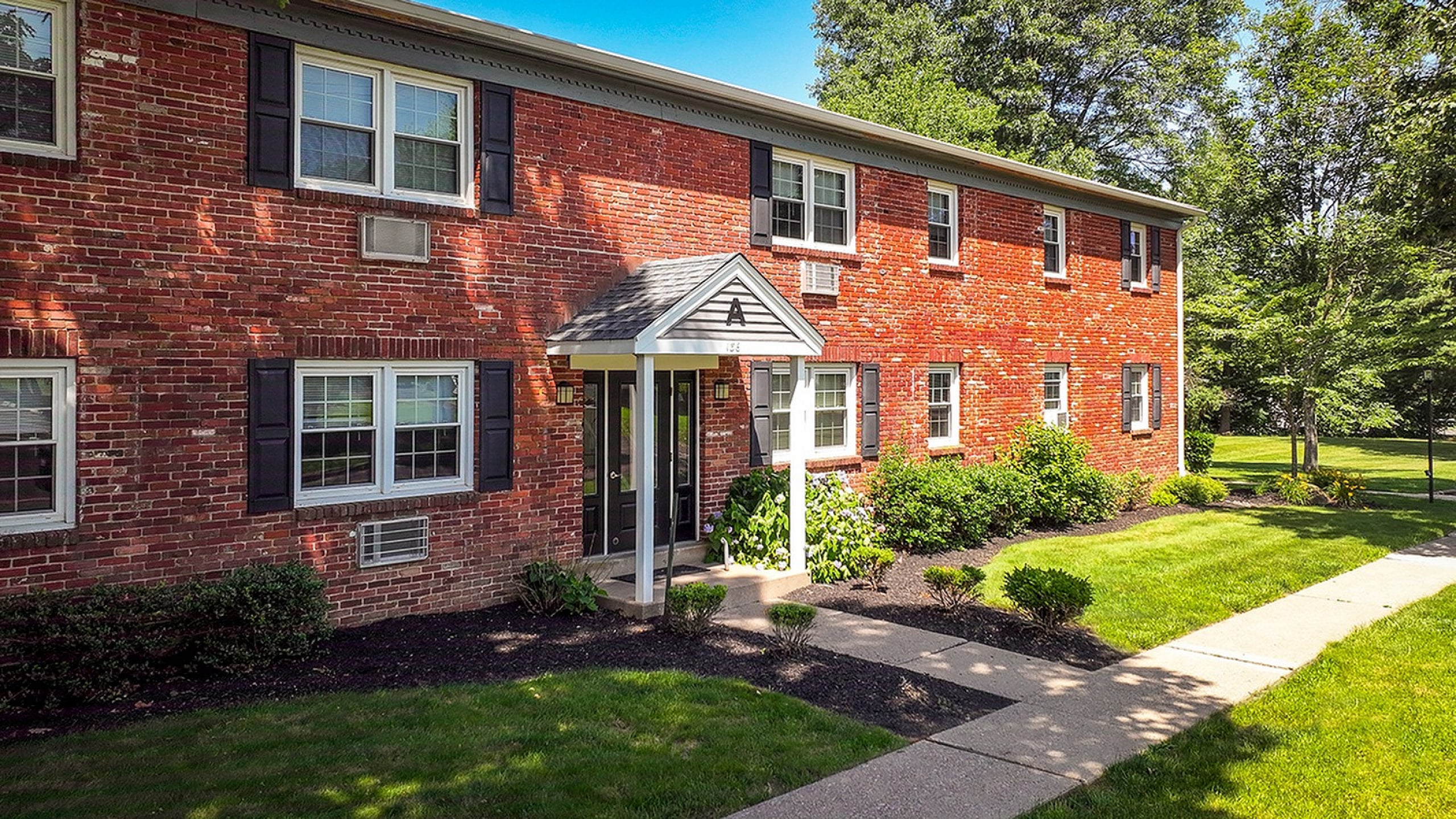 Charming brick apartment building surrounded by lush greenery and manicured lawns, featuring a welcoming entrance with classic black shutters and vibrant landscaping.