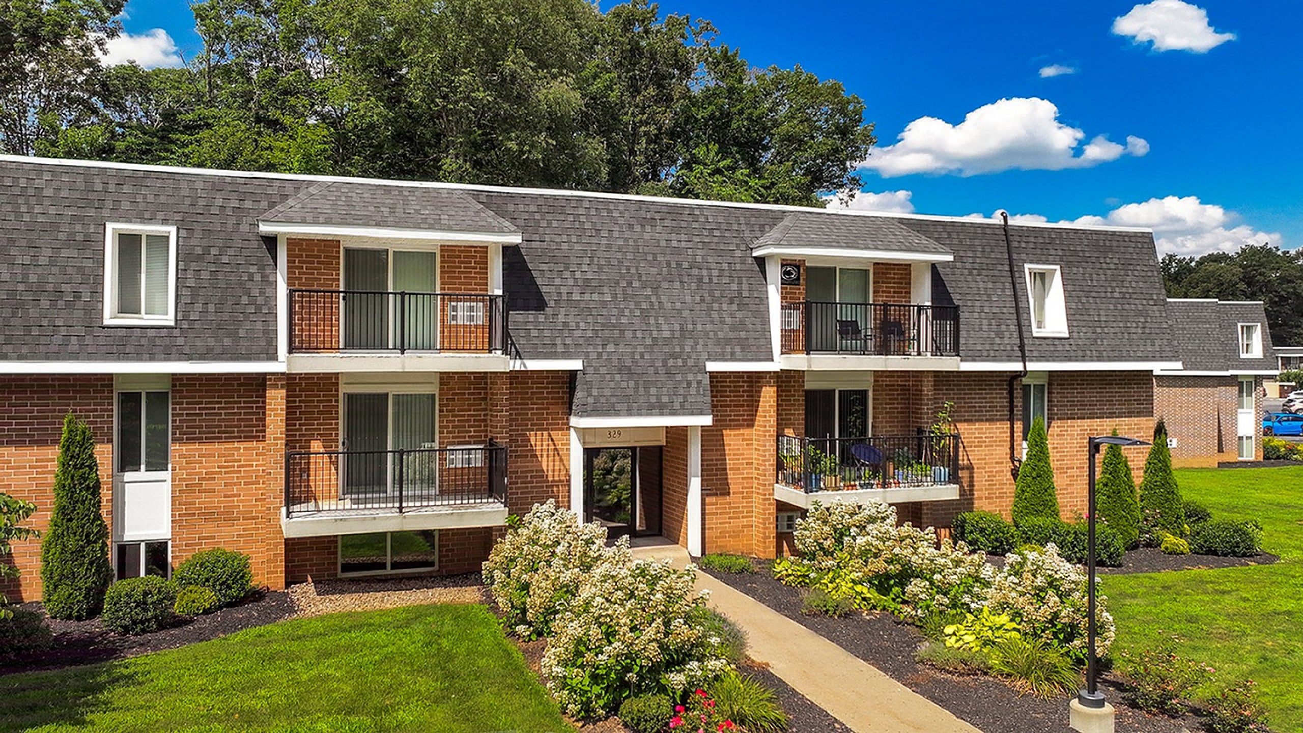 Charming two-story brick apartment building with private balconies surrounded by lush greenery and beautiful landscaping under a vibrant blue sky.