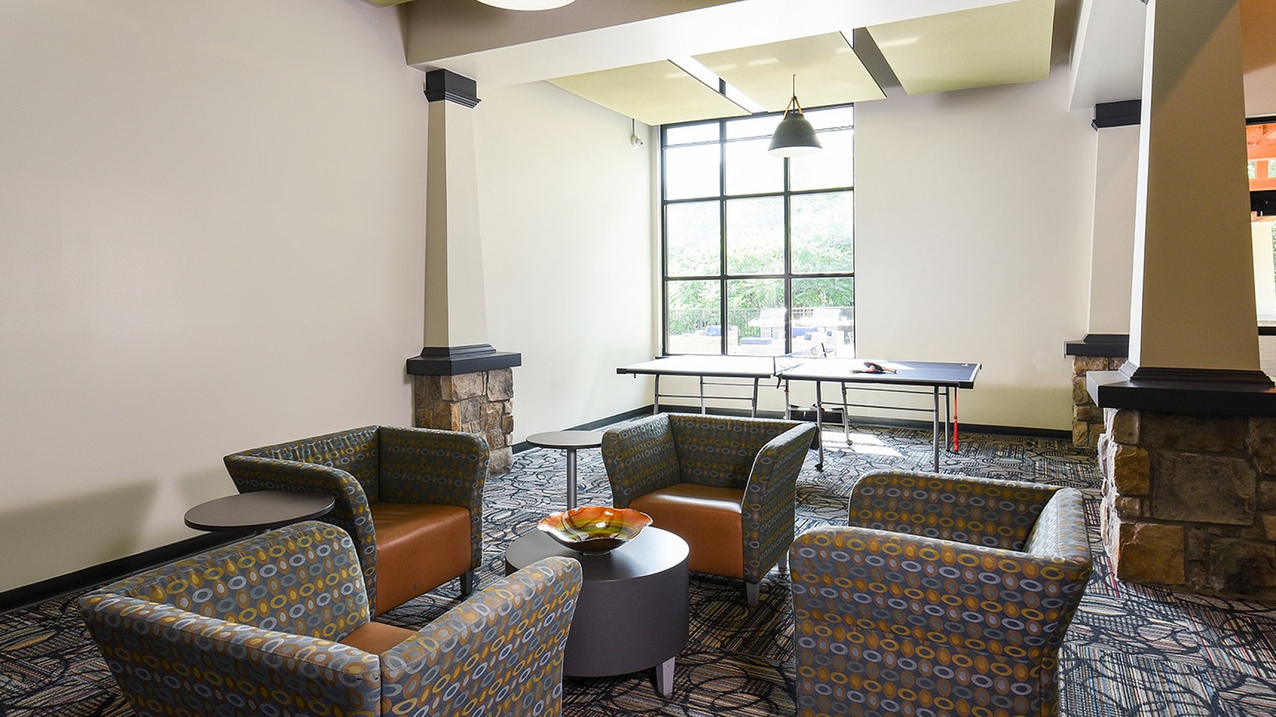 Modern communal lounge with stylish seating, accent lighting, and a ping pong table by a large window.