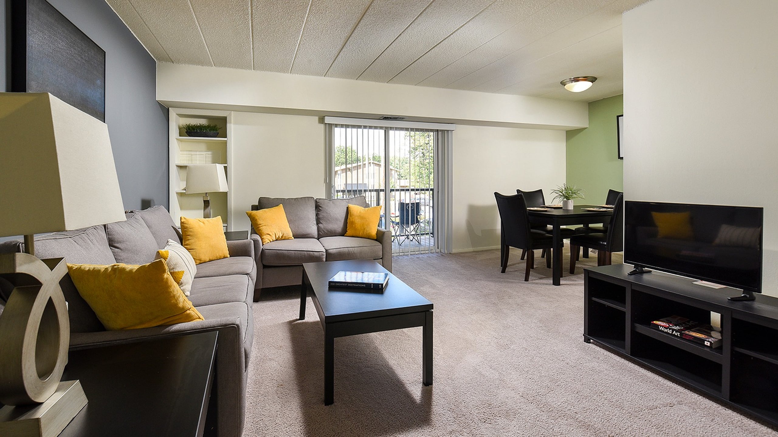 Inviting living room with plush gray sofas, vibrant yellow accent pillows, and a sleek modern design, featuring a cozy dining area and large sliding glass doors leading to a bright outdoor space.