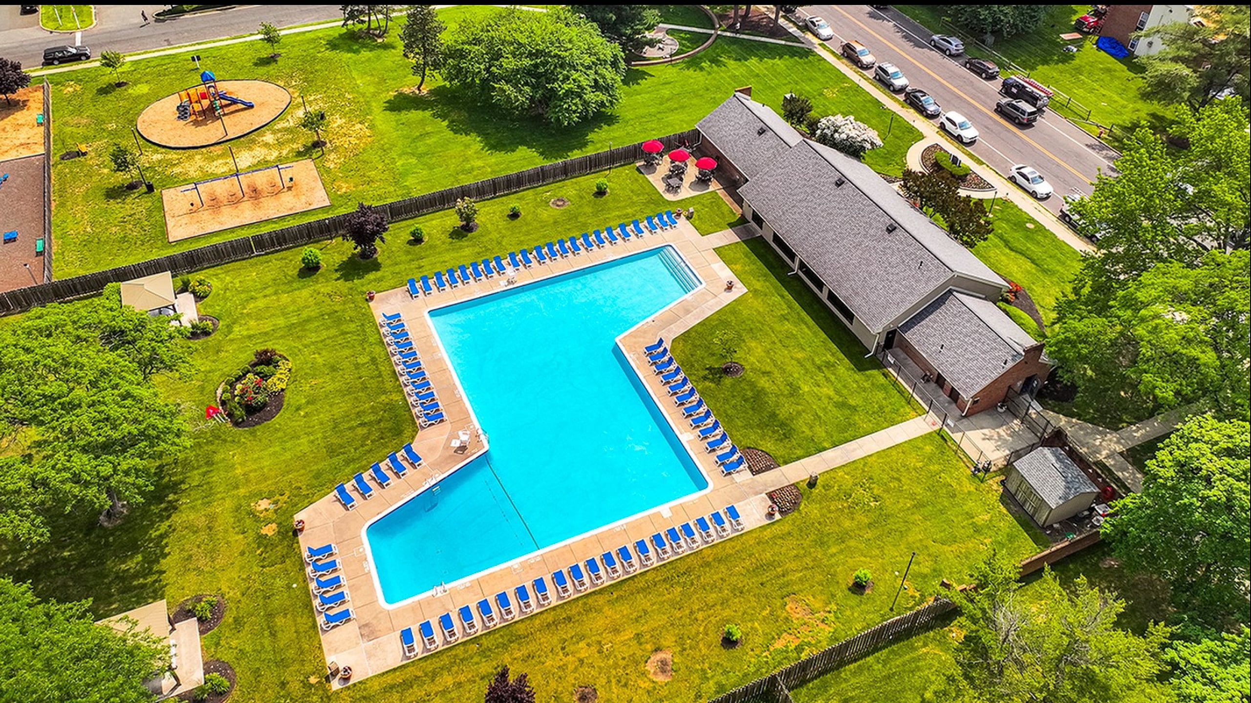 Aerial view of a luxurious community pool area with numerous blue sun loungers, surrounded by lush green lawns and landscaped gardens, adjacent to a charming clubhouse and a family-friendly playground.