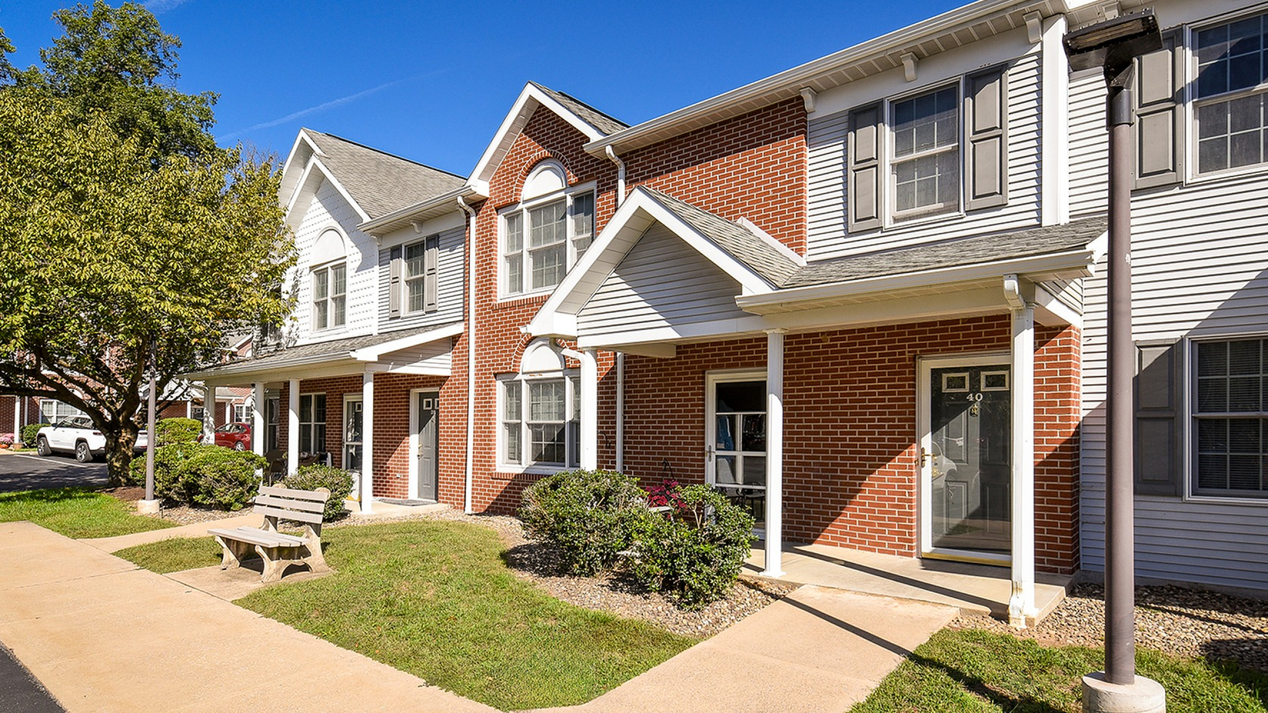 Charming townhouse exterior with classic red brick facade, lush green landscaping, and inviting front porch under a clear blue sky.
