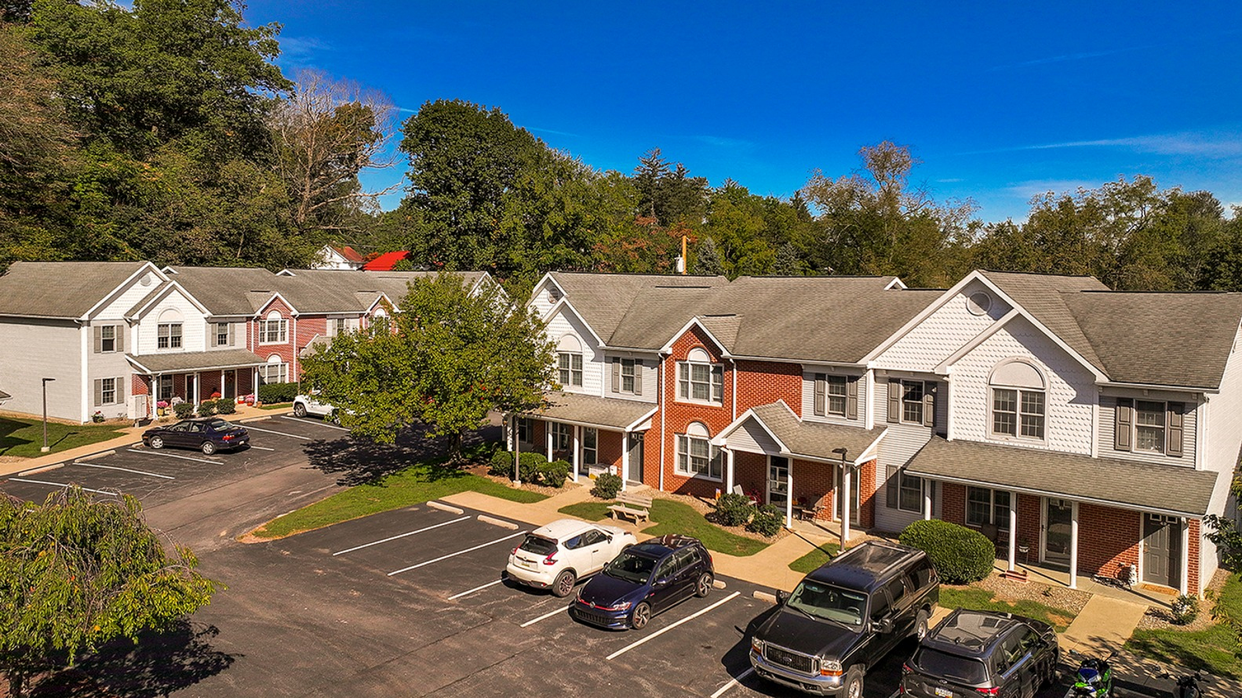 Charming suburban townhouses with welcoming porches and ample parking, surrounded by lush greenery and a vibrant blue sky.