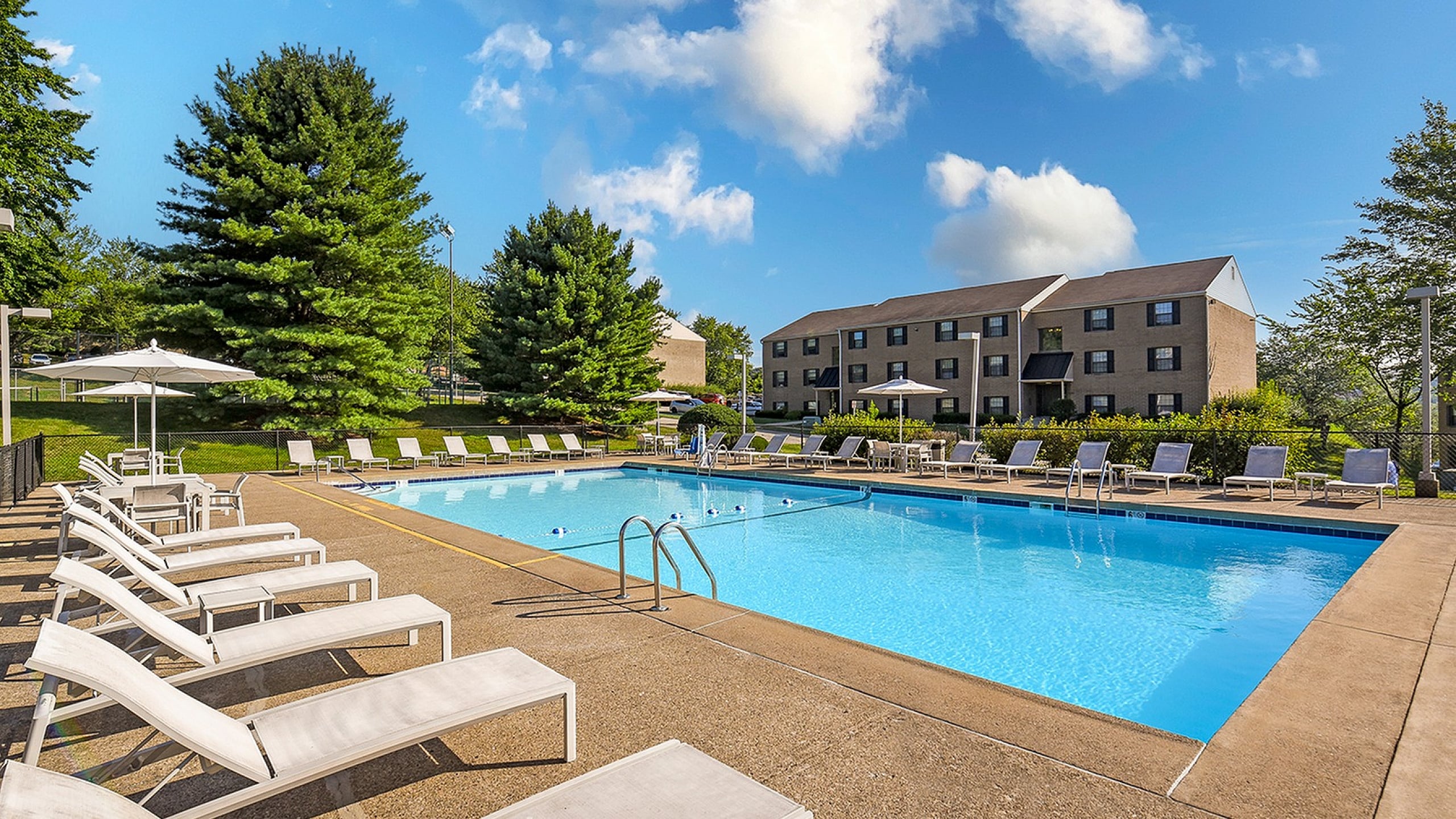 Inviting outdoor pool with sun loungers and umbrellas, surrounded by lush greenery and modern apartment buildings under a clear blue sky.