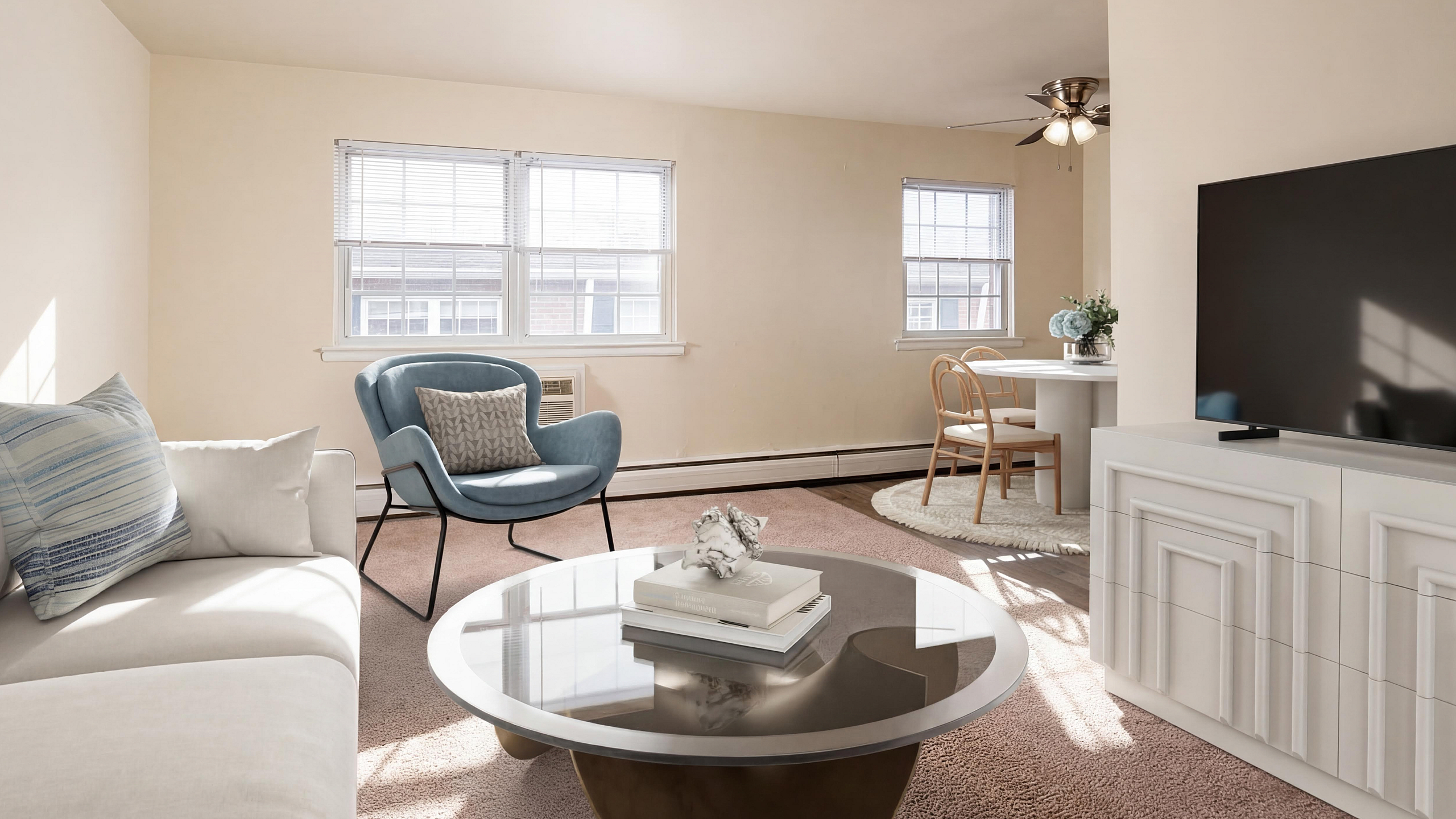 Bright, inviting living room with a cozy sofa, stylish blue armchair, and elegant round coffee table. Adjacent dining area featuring chic wooden chairs and a modern table. Sunlight streams through large windows, creating a warm, welcoming a