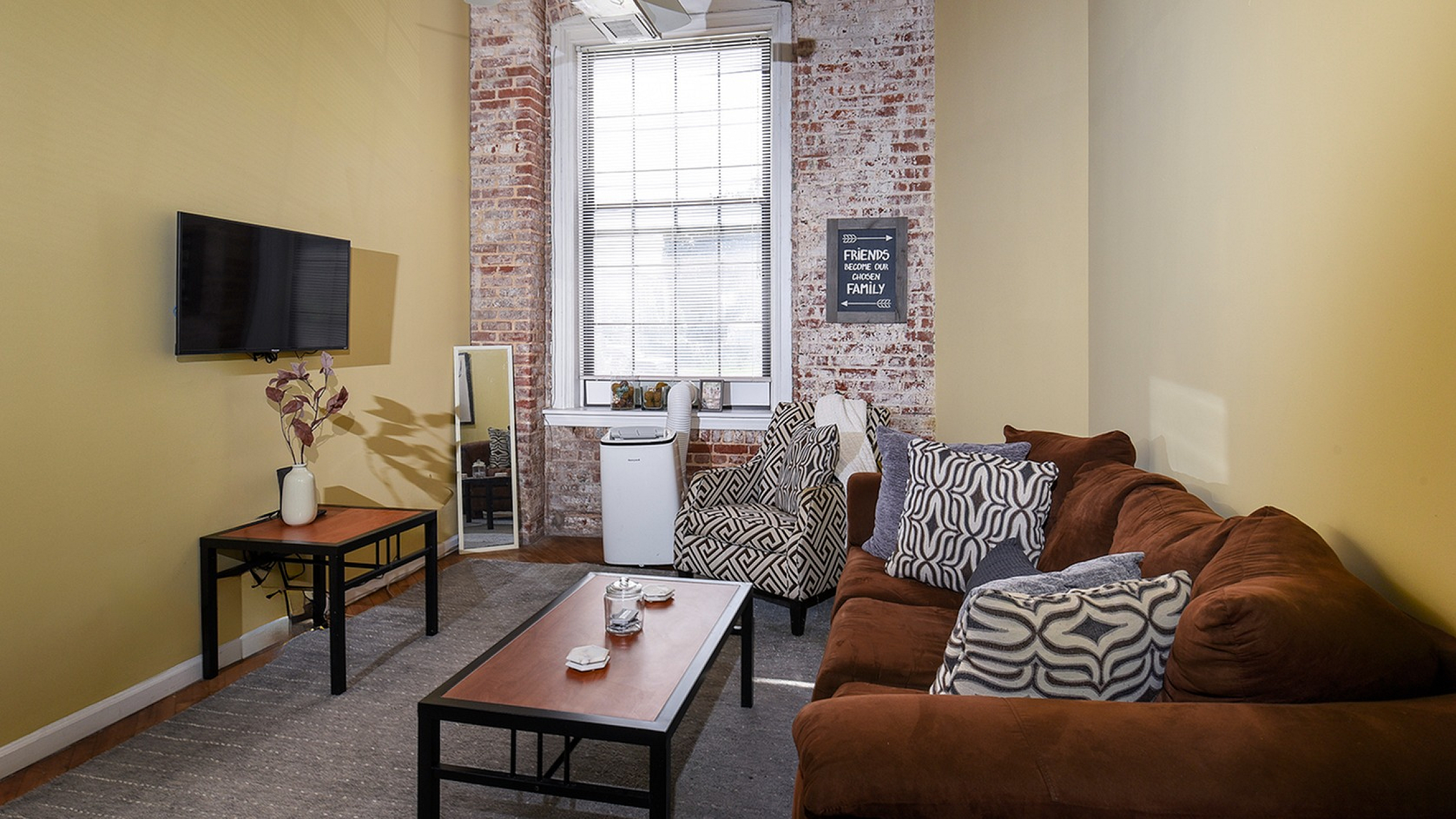 Cozy living room with exposed brick walls, large window, stylish brown sofa, and a flat-screen TV for modern urban living.