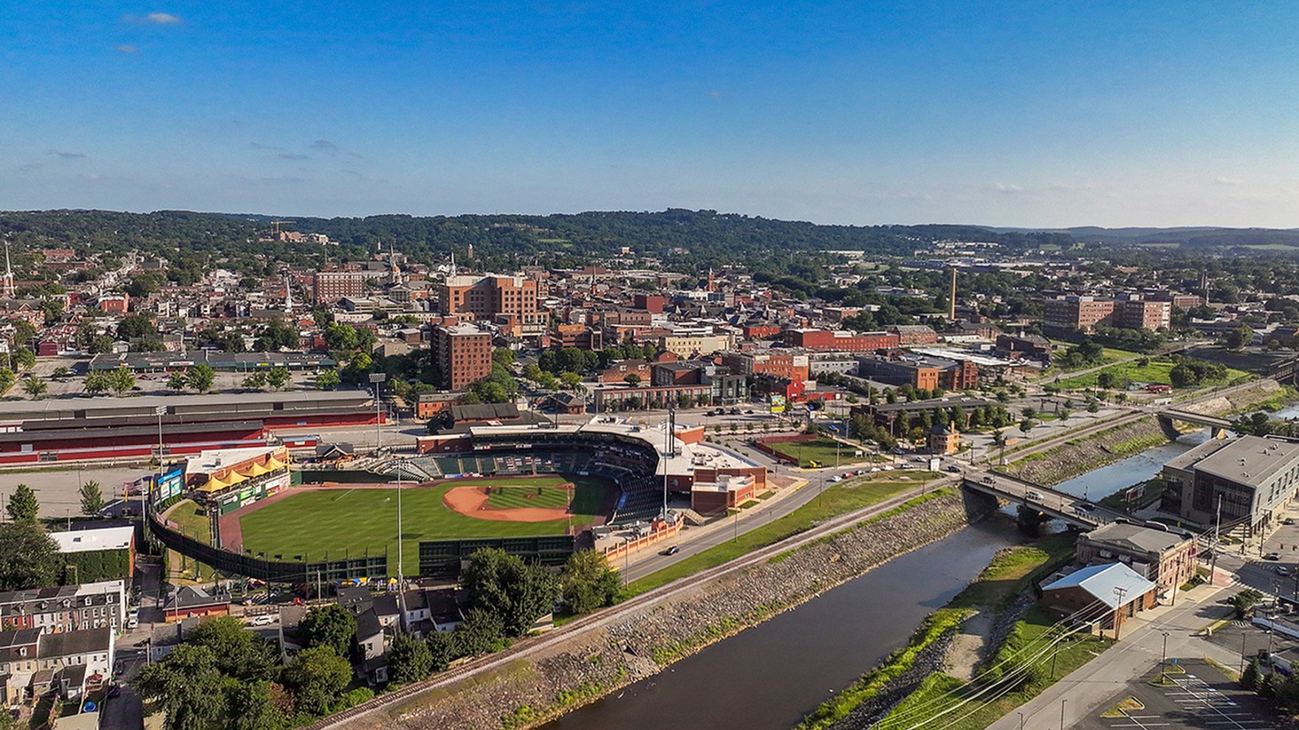 Vibrant aerial view of a charming cityscape featuring a picturesque baseball stadium, surrounded by lush greenery and historic architecture, with a serene river meandering through the town.