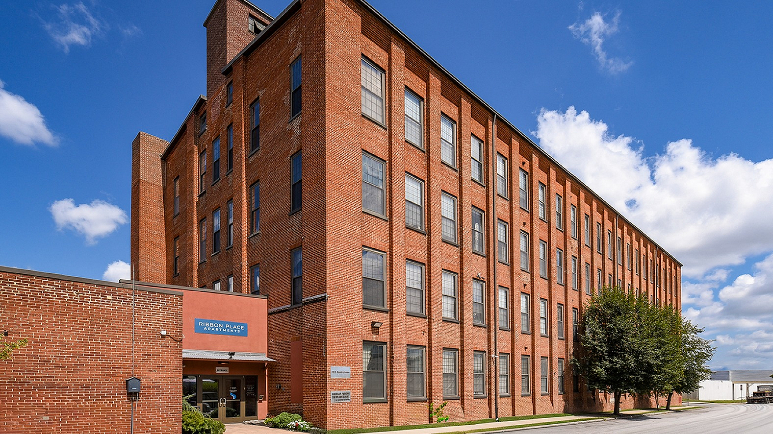 Charming brick apartment building with a classic industrial design, featuring large windows and lush greenery, set against a bright blue sky.