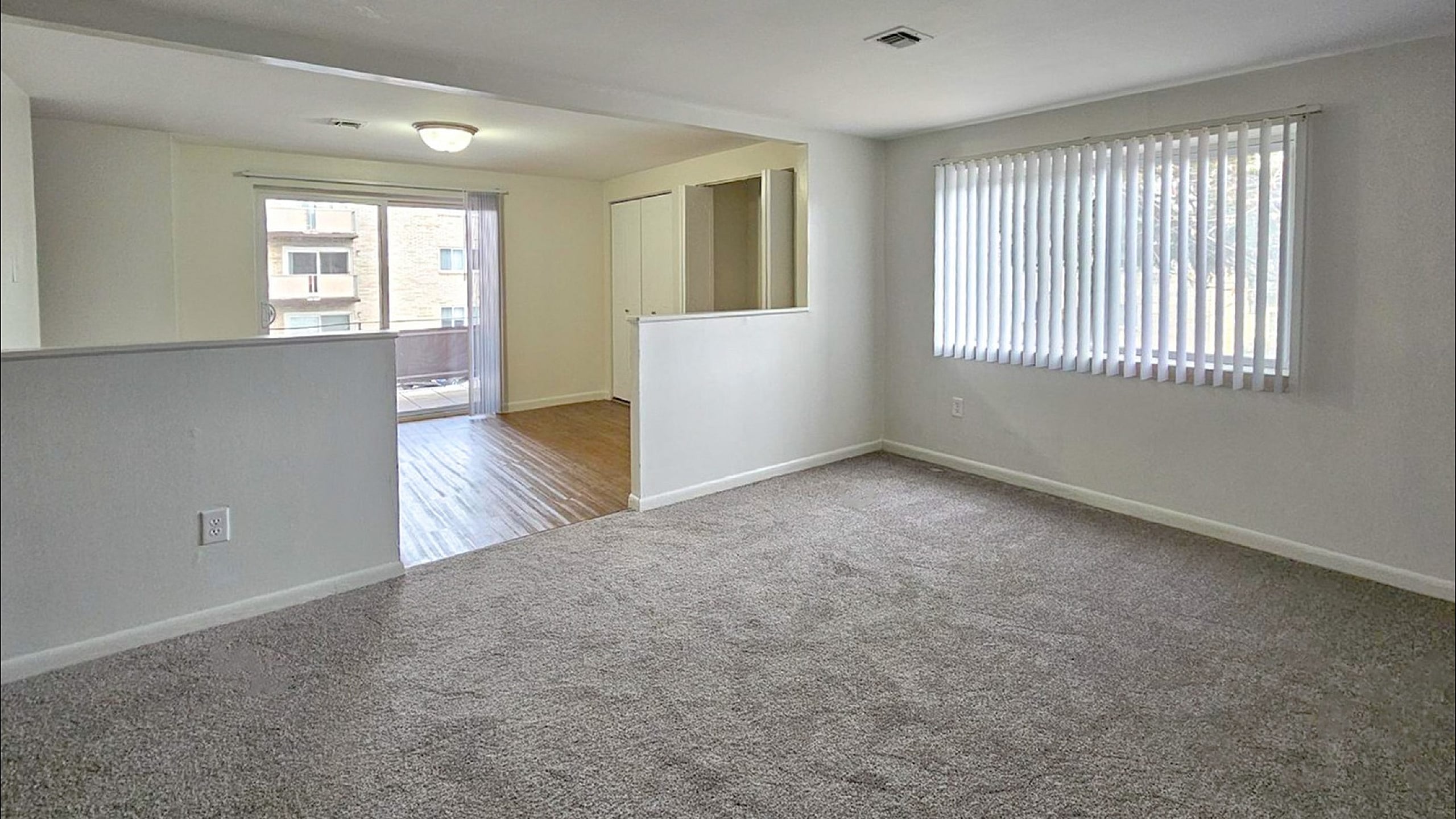 Bright and spacious living area with large windows, carpet flooring, and open flow to a hardwood space, ideal for seamless living.