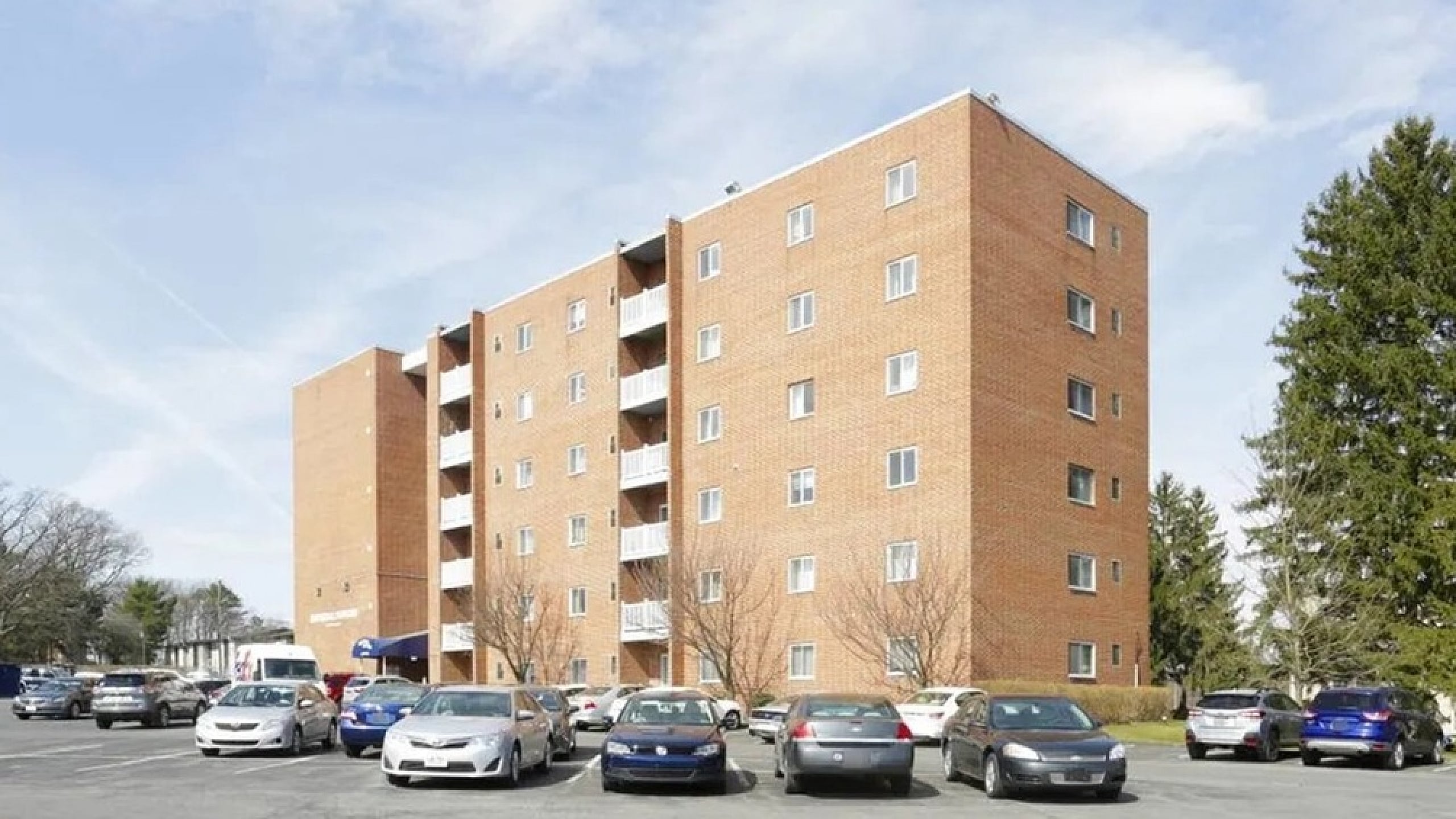 Charming brick apartment building with spacious parking area, surrounded by greenery and blue skies.