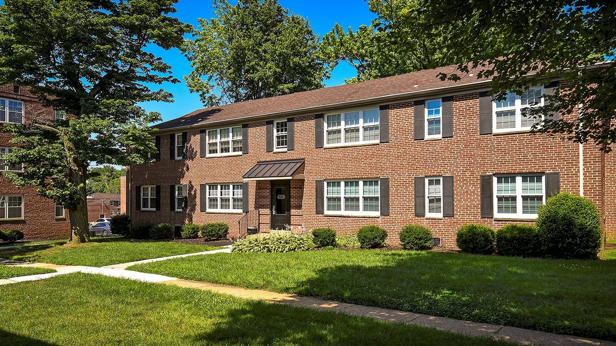 Charming brick apartment building surrounded by lush green lawns and mature trees, basking in sunlight for a serene residential experience.