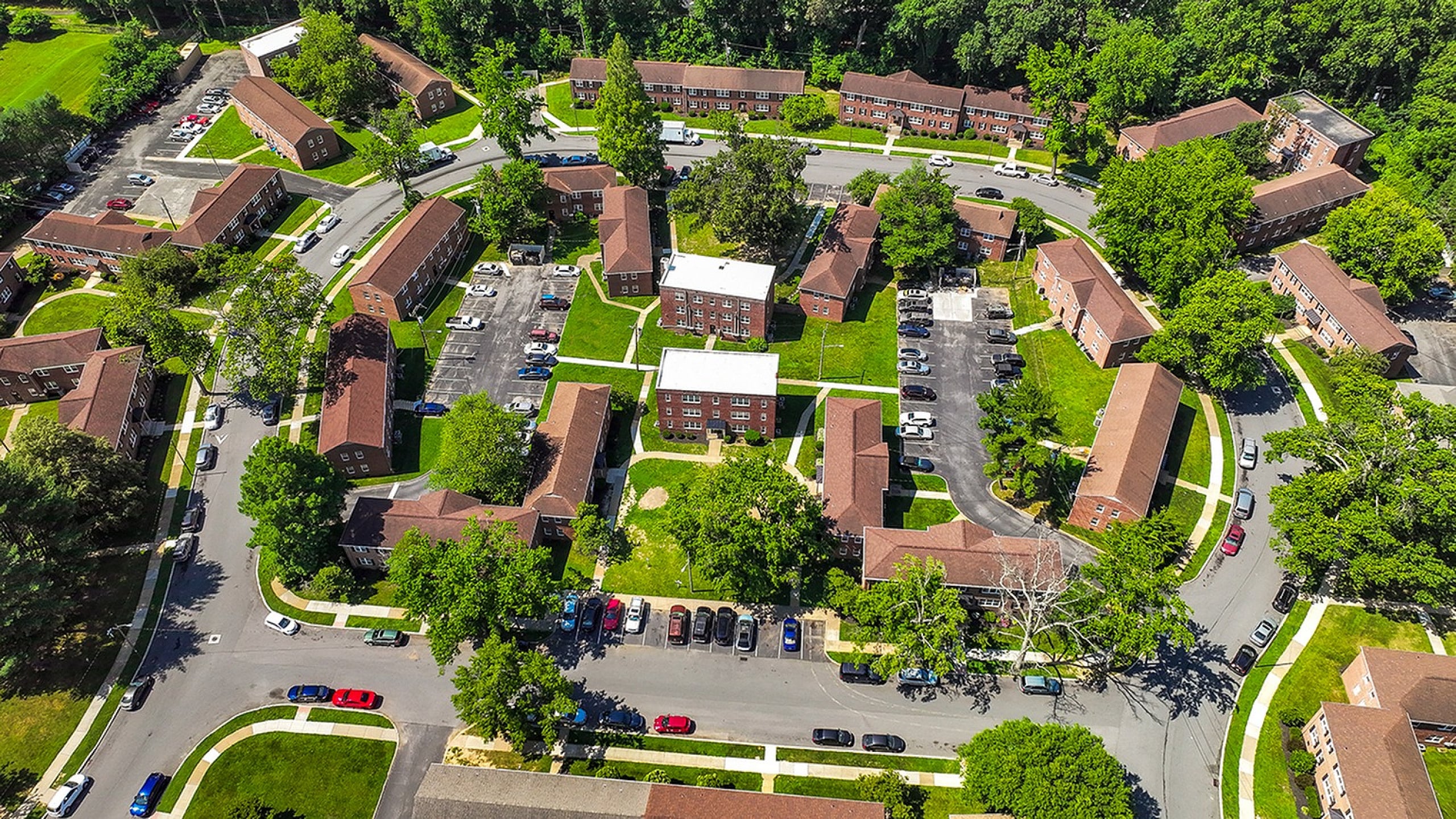 Vibrant aerial view of a charming apartment complex surrounded by lush greenery and ample parking spaces, offering a serene community living experience.
