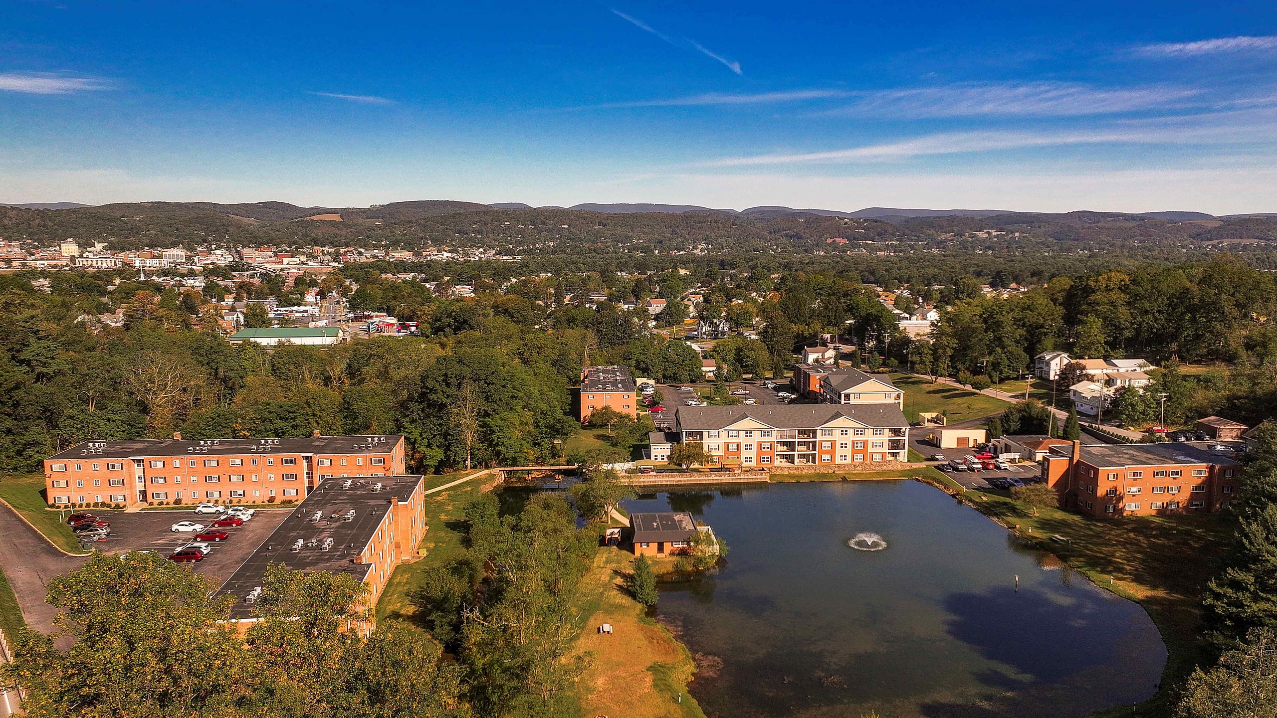 Charming aerial view of a residential community nestled amidst lush greenery, featuring elegant brick buildings and a serene pond with a fountain.