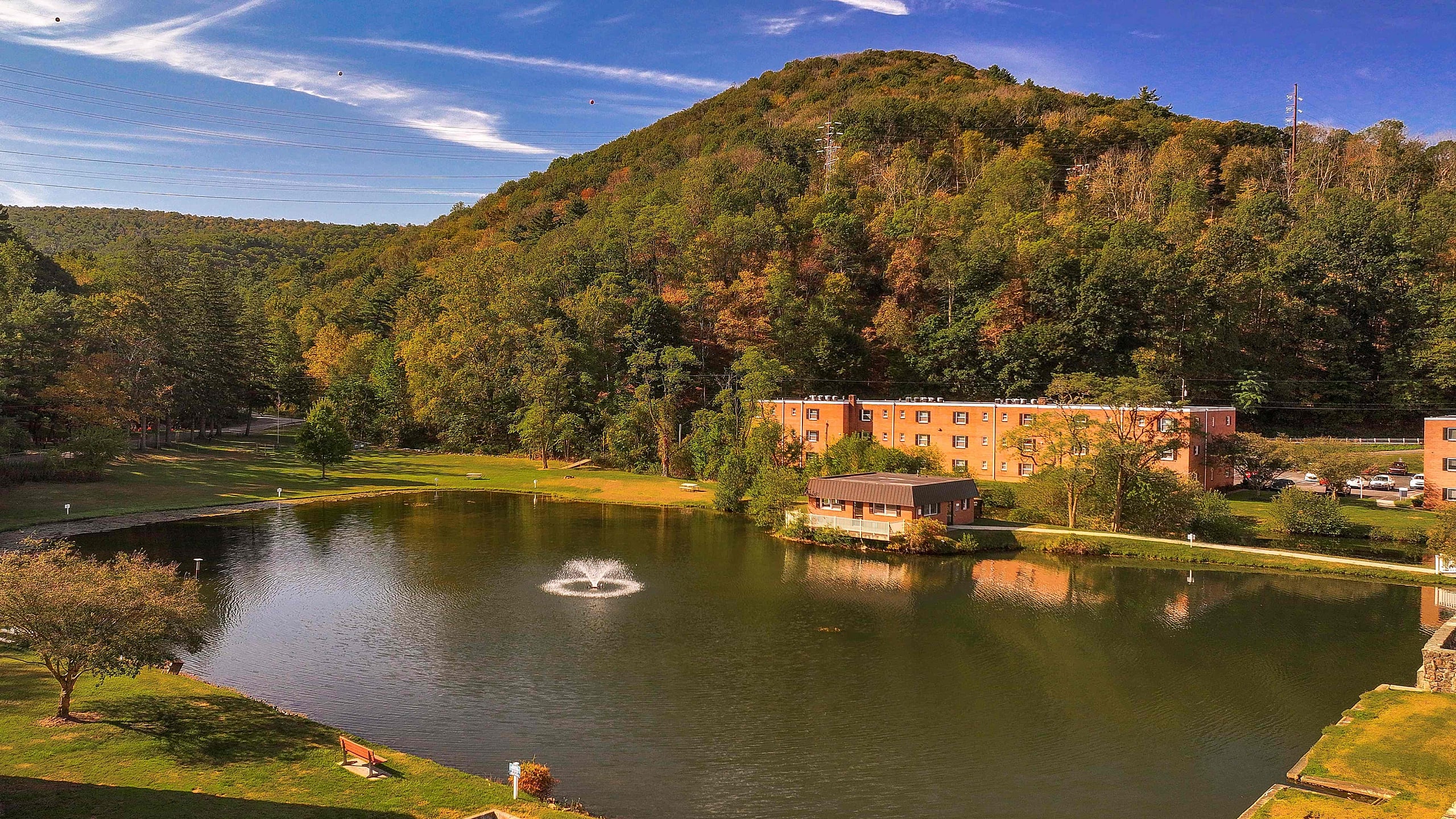 Scenic view of serene pond with a fountain, surrounded by lush trees and a charming apartment building nestled against a picturesque hillside.