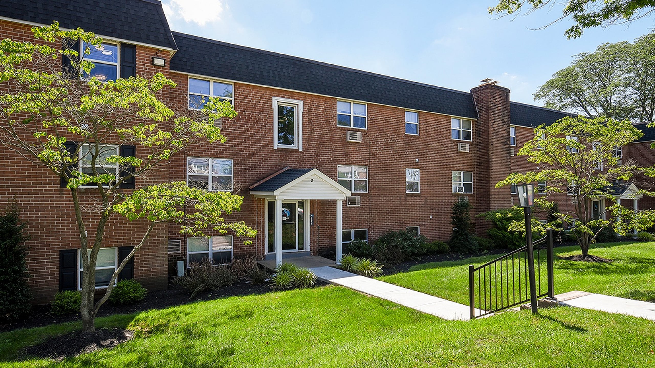 Charming brick apartment building surrounded by lush greenery and well-kept lawns, reflecting a welcoming and serene atmosphere.