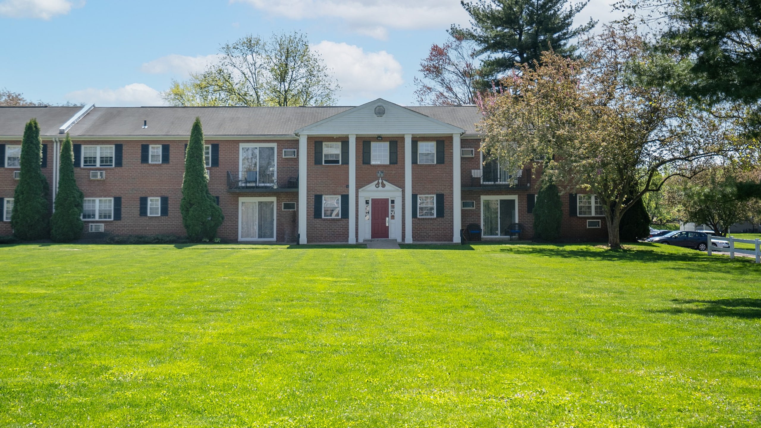 Charming brick apartment building nestled in lush green surroundings, featuring classic architecture with white columns and inviting red doors.