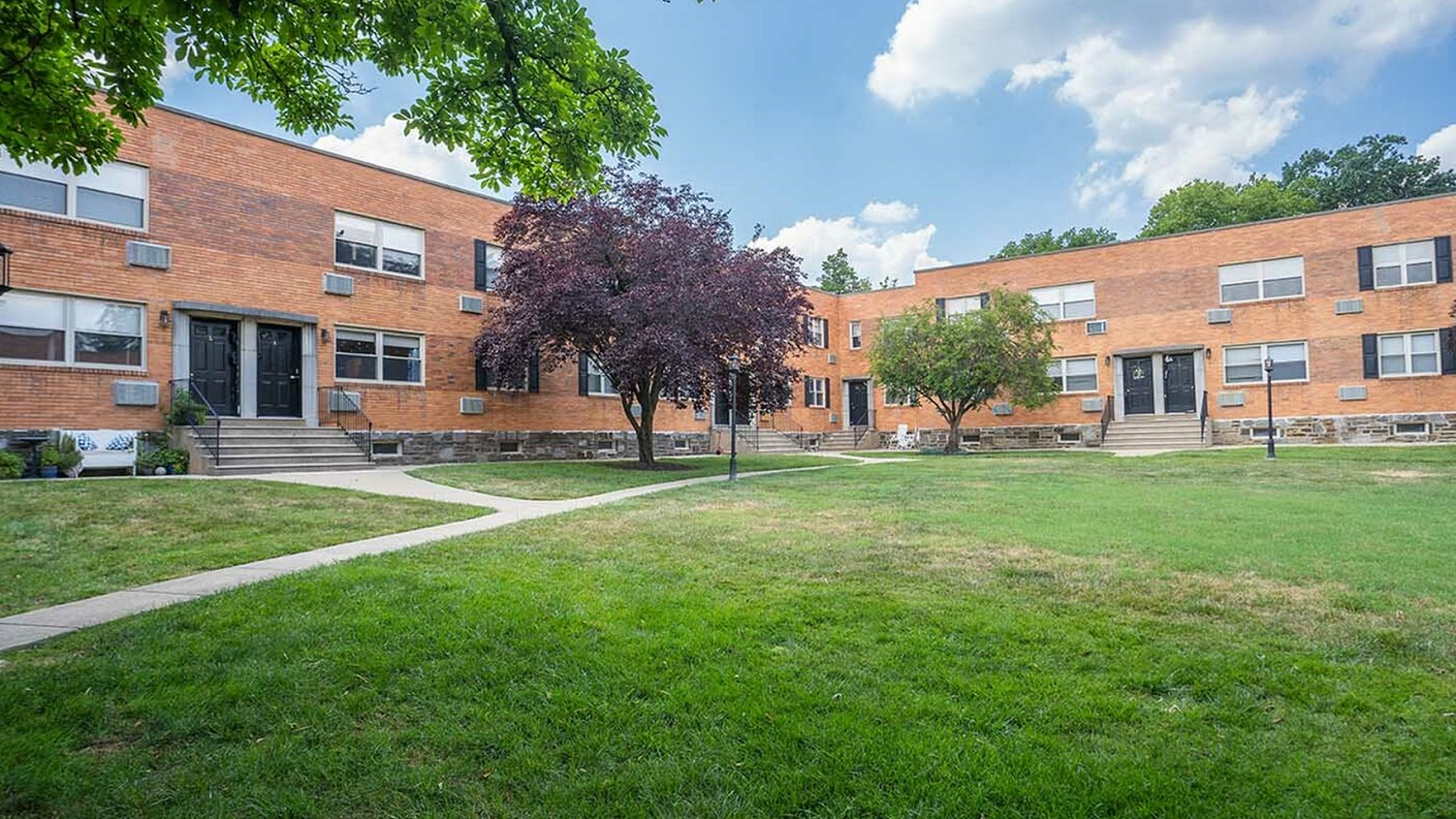 Inviting apartment complex with charming brick facade, lush green lawn, and mature trees under a clear blue sky.