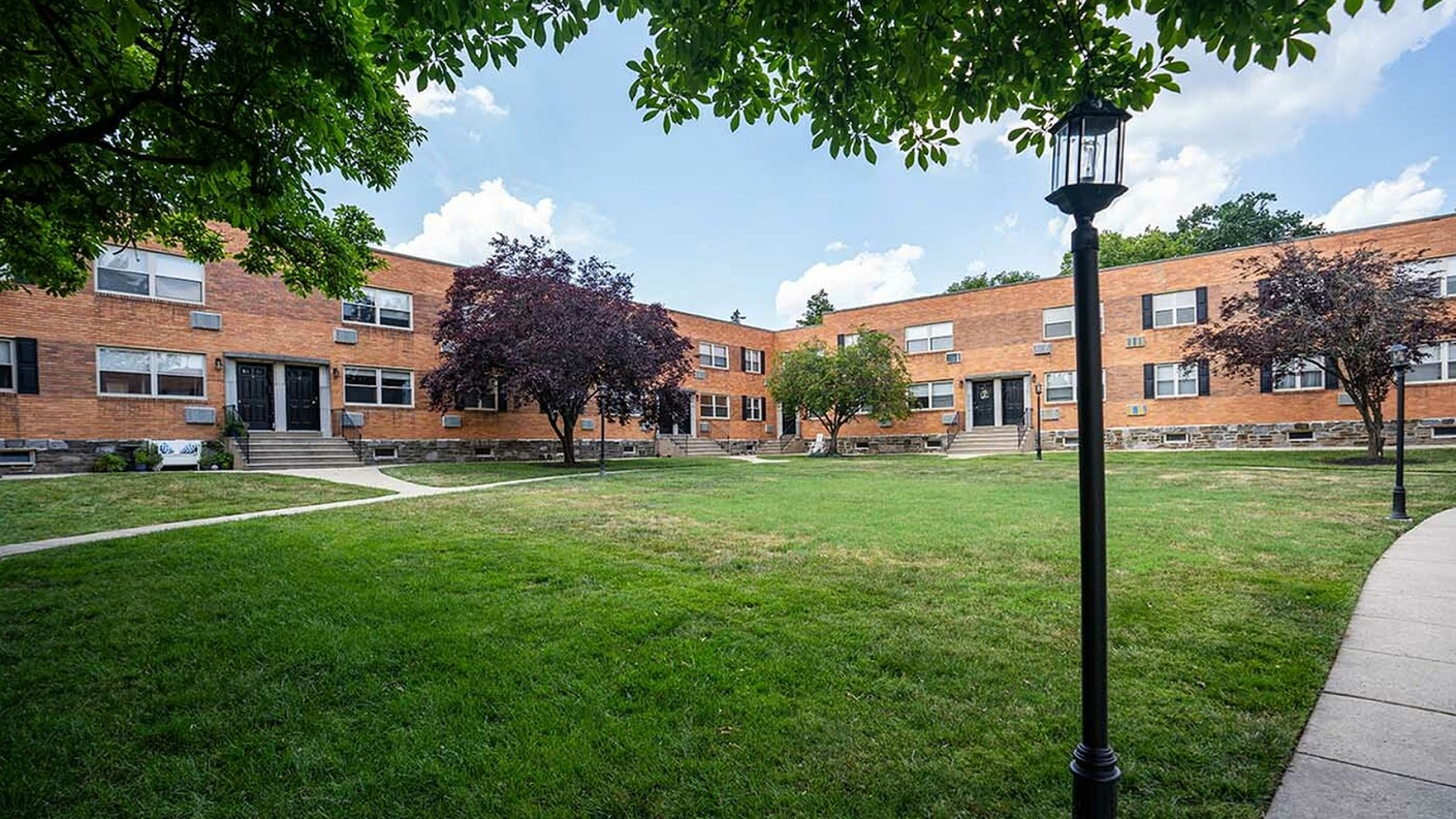 Charming brick apartment building with manicured lawns, surrounded by mature trees and inviting pathways.