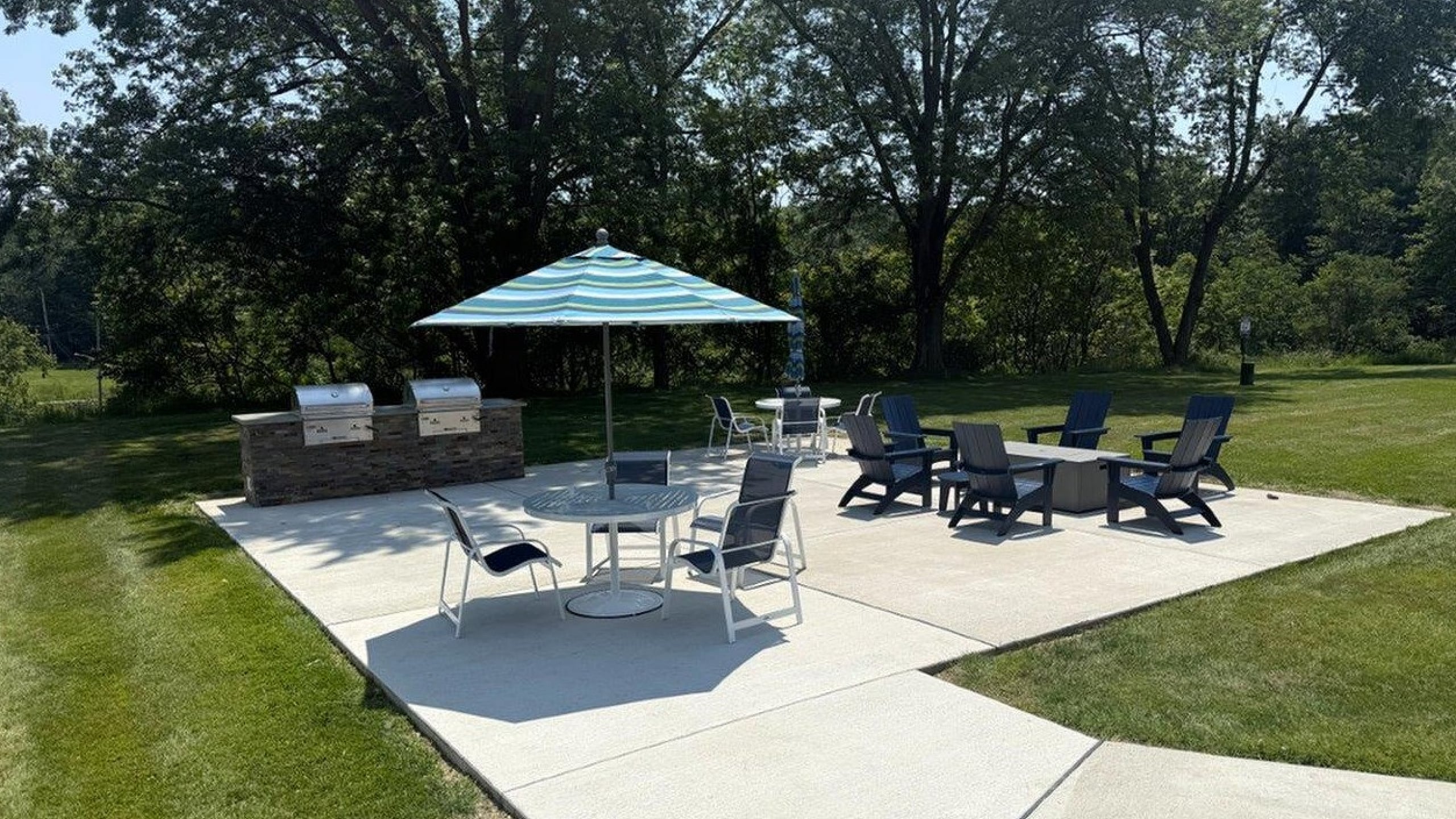 Modern outdoor patio with stylish seating, BBQ grills, and large umbrella, perfect for entertaining in a lush green setting.