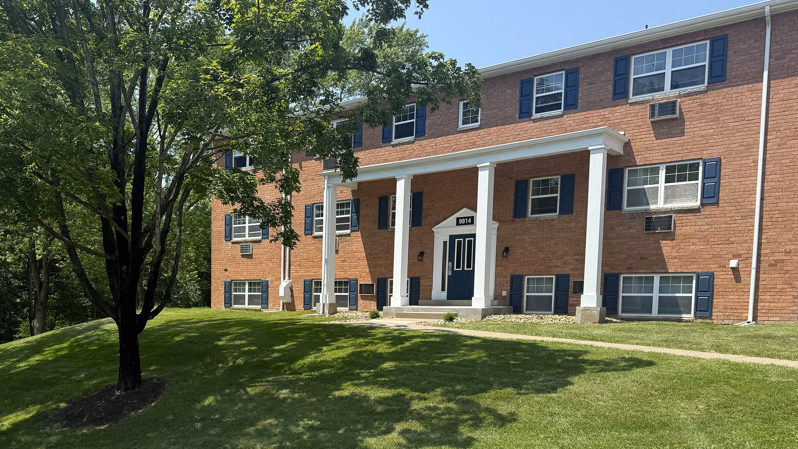 Charming brick apartment building with elegant columns, surrounded by lush green lawns and mature trees on a sunny day.